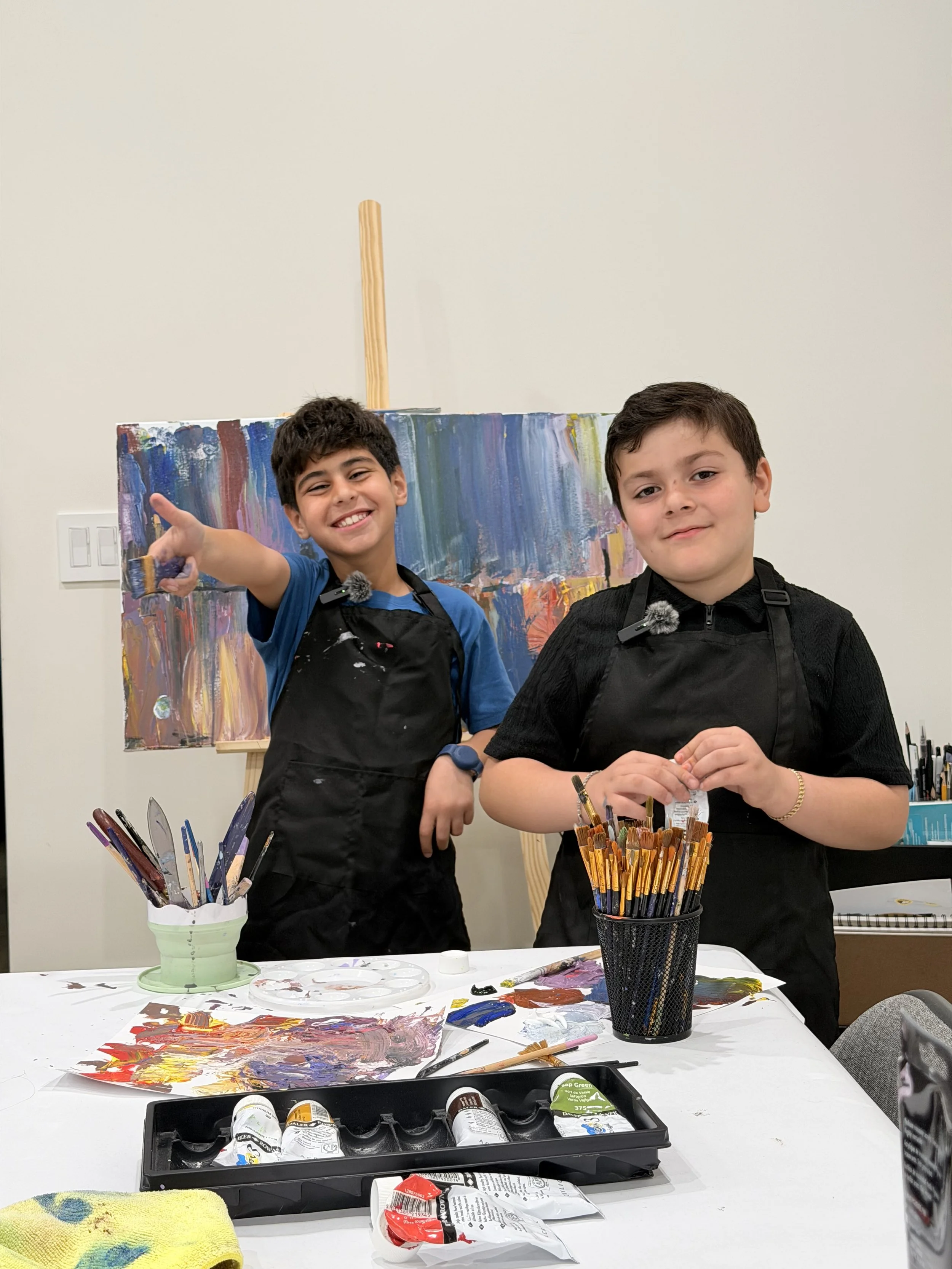 Two young boys are standing behind a table filled with painting supplies, including paintbrushes, paints, and paints tubes. The boy on the left is smiling and pointing at something outside the frame, wearing a blue shirt and a black apron. The boy on