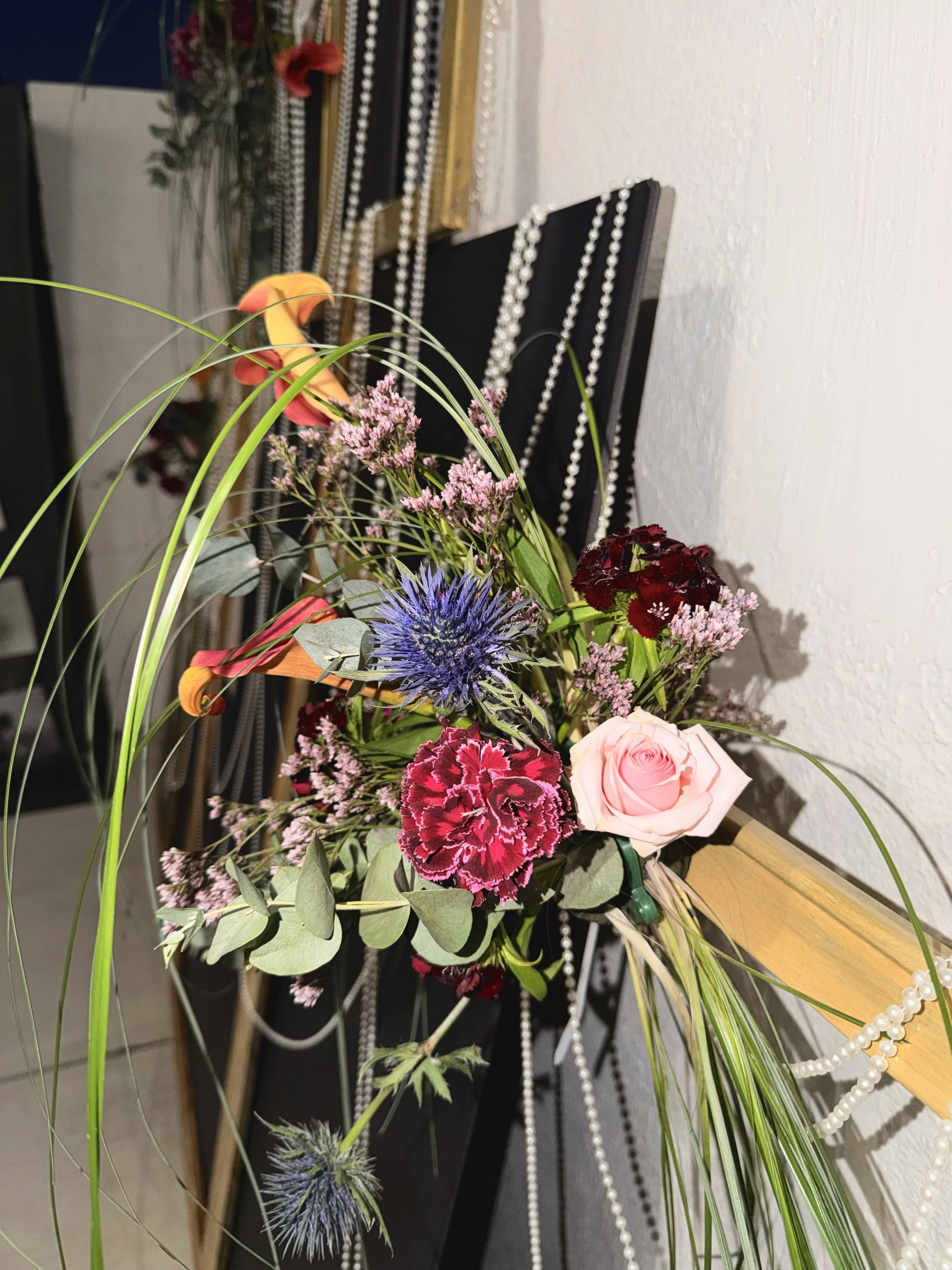 A floral arrangement with pink roses, purple thistle, red and pink carnations, orange lilies, and green foliage, placed in front of a black display with pearl necklaces hanging on it.