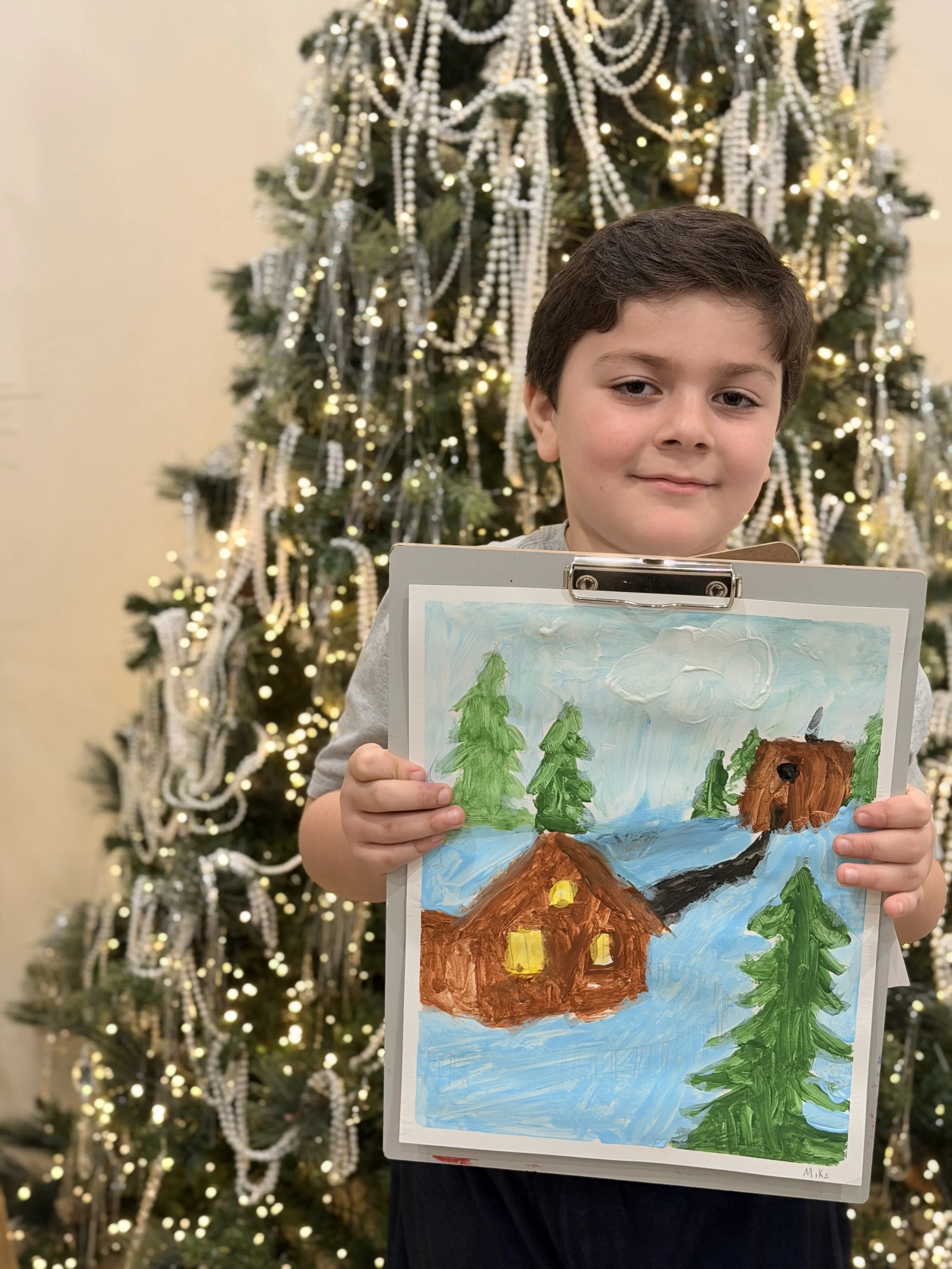 A boy with brown hair holding a painted picture of a landscape scene with green trees, a brown bear, and blue sky in front of a decorated Christmas tree with white pearls and warm lights.