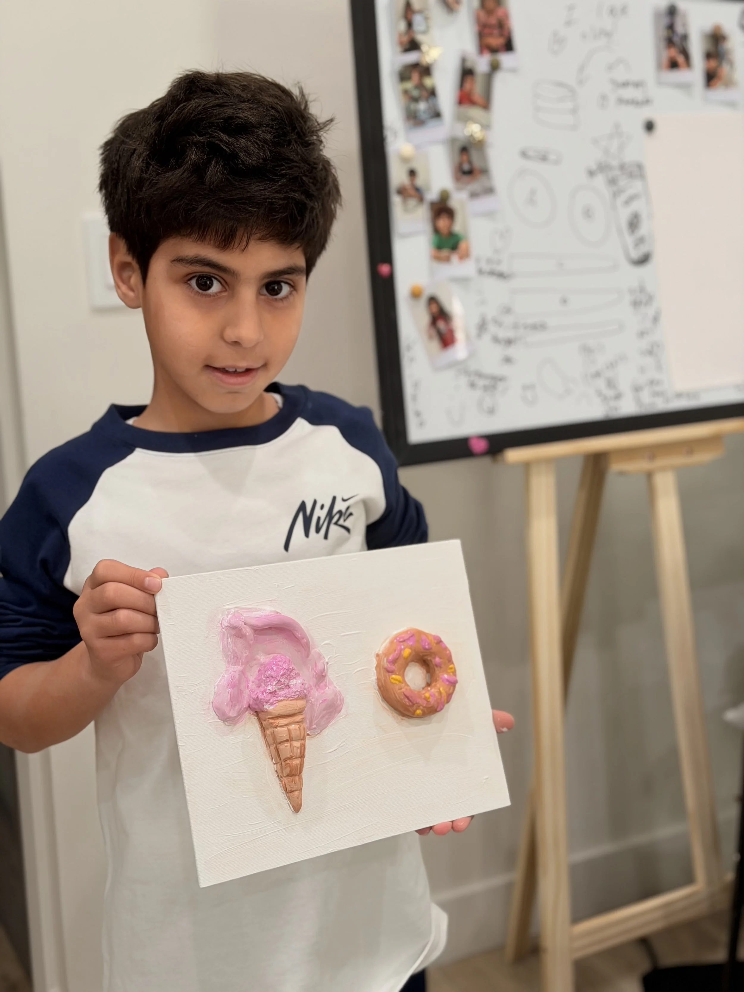 A young boy holding an art project of a pink ice cream cone and a pink donut on a white canvas, standing in front of a whiteboard with photos and drawings.