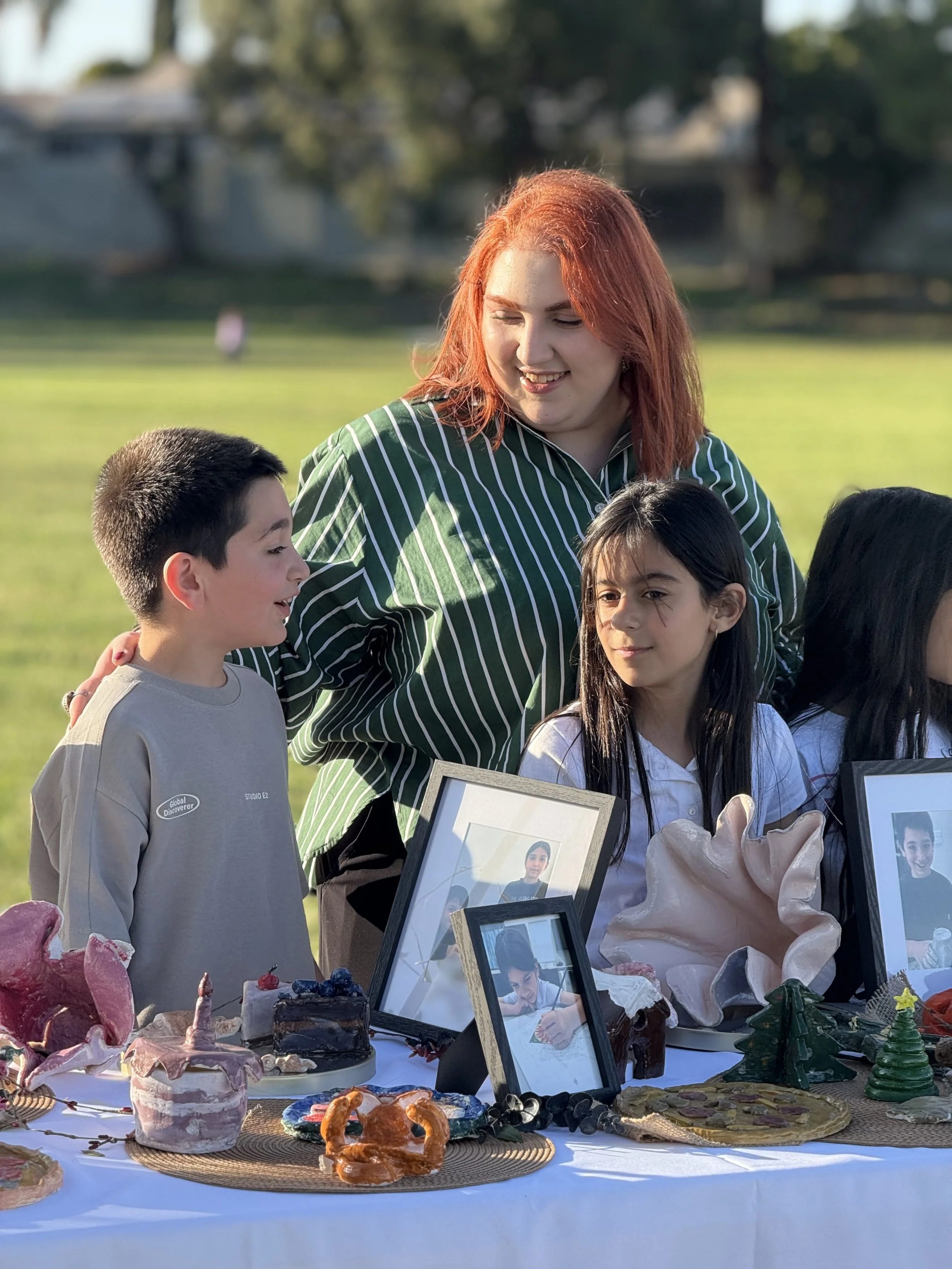 A woman with red hair and three children standing behind a table filled with baked goods and framed photos. The scene is outdoors on a sunny day.