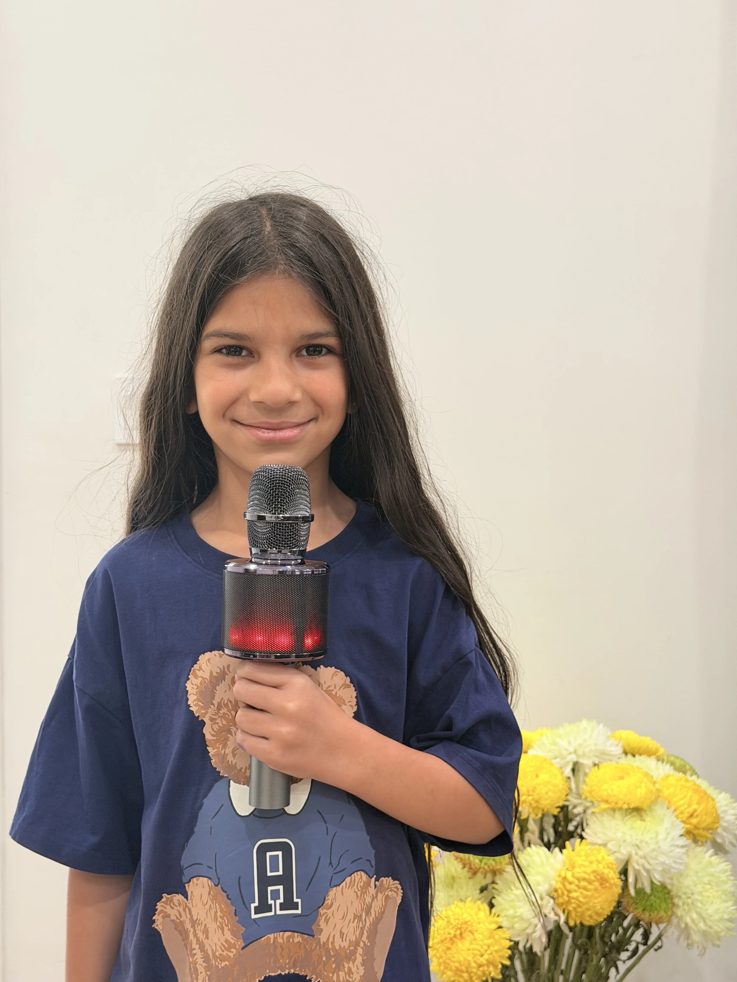 A young girl with long dark hair smiling while holding a microphone, standing next to a large bouquet of white and yellow flowers against a plain light-colored wall.