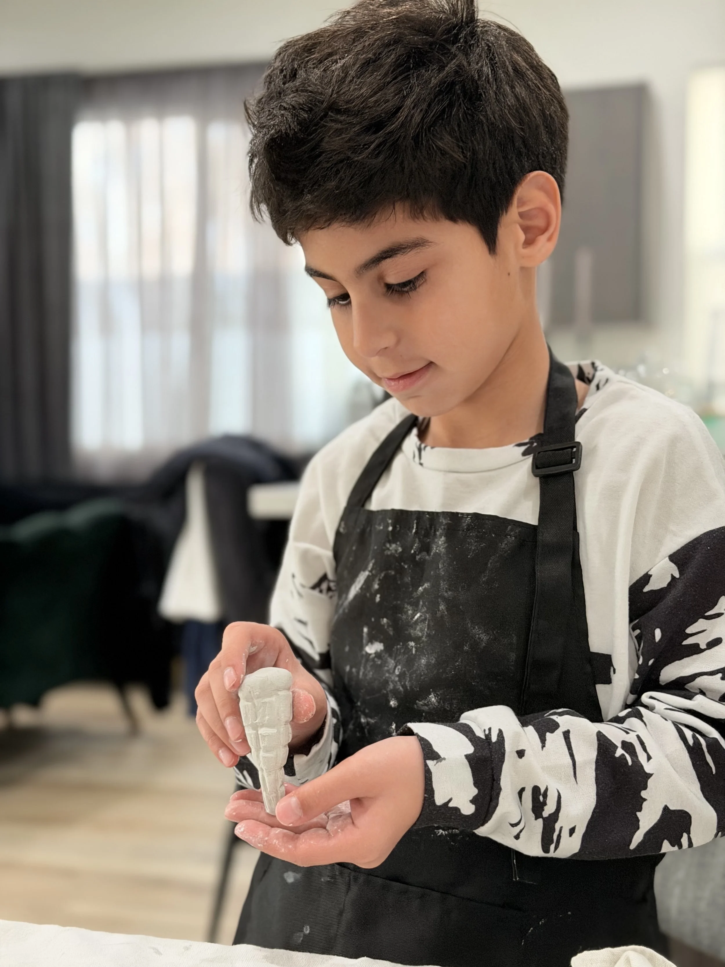 A young boy with dark hair and a patterned shirt, wearing a black apron, is working on a sculpture with clay, shaping it with his hands in a well-lit indoor space.