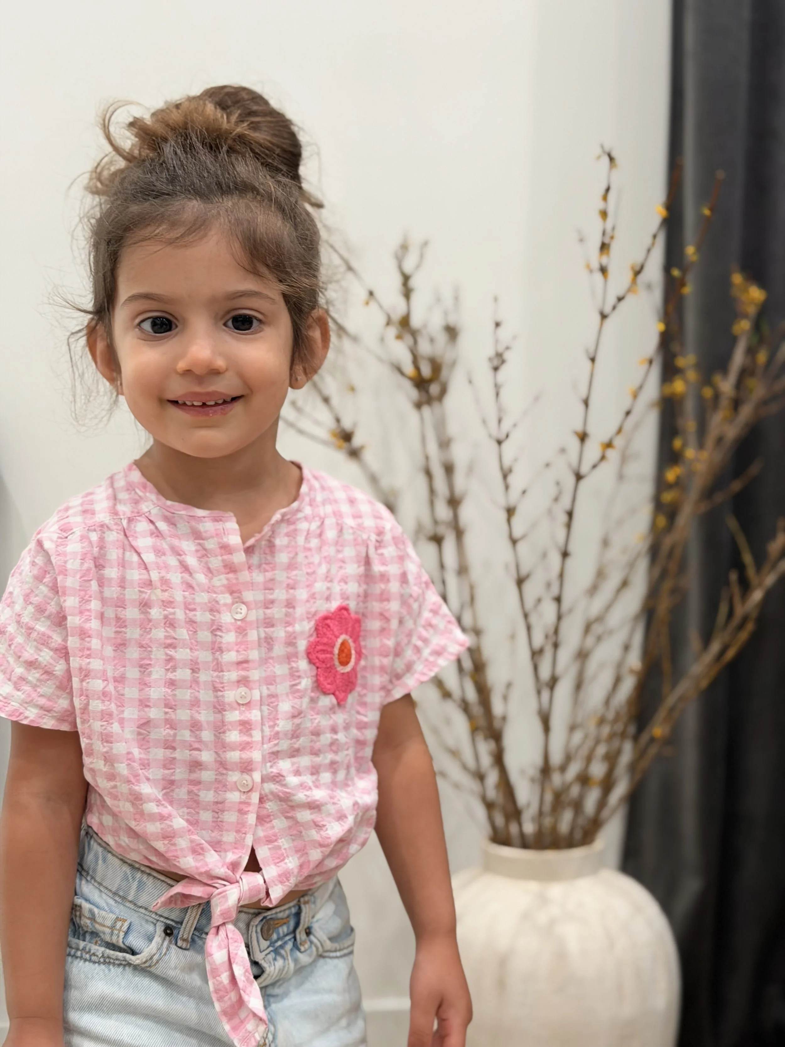 A young girl with brown hair tied up in a bun, wearing a pink checkered shirt with a flower applique, standing in front of a white wall and a large vase with branches.