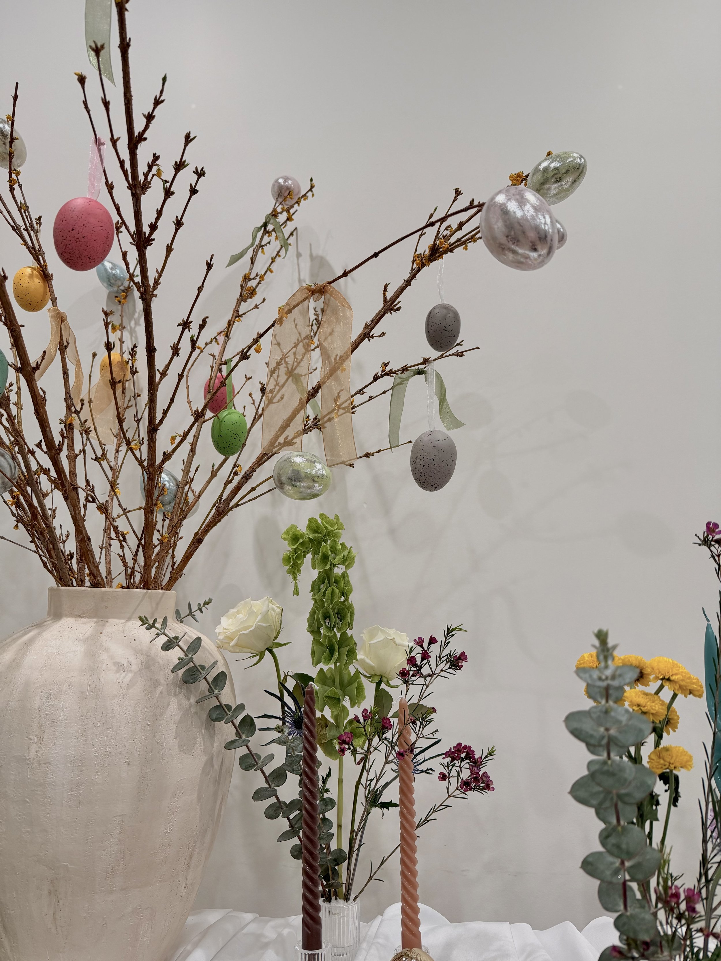 Vase with small tree branches decorated with pastel-colored and speckled egg ornaments, ribbons, and greenery, placed on a table with white tablecloth, Easter, against a plain wall.