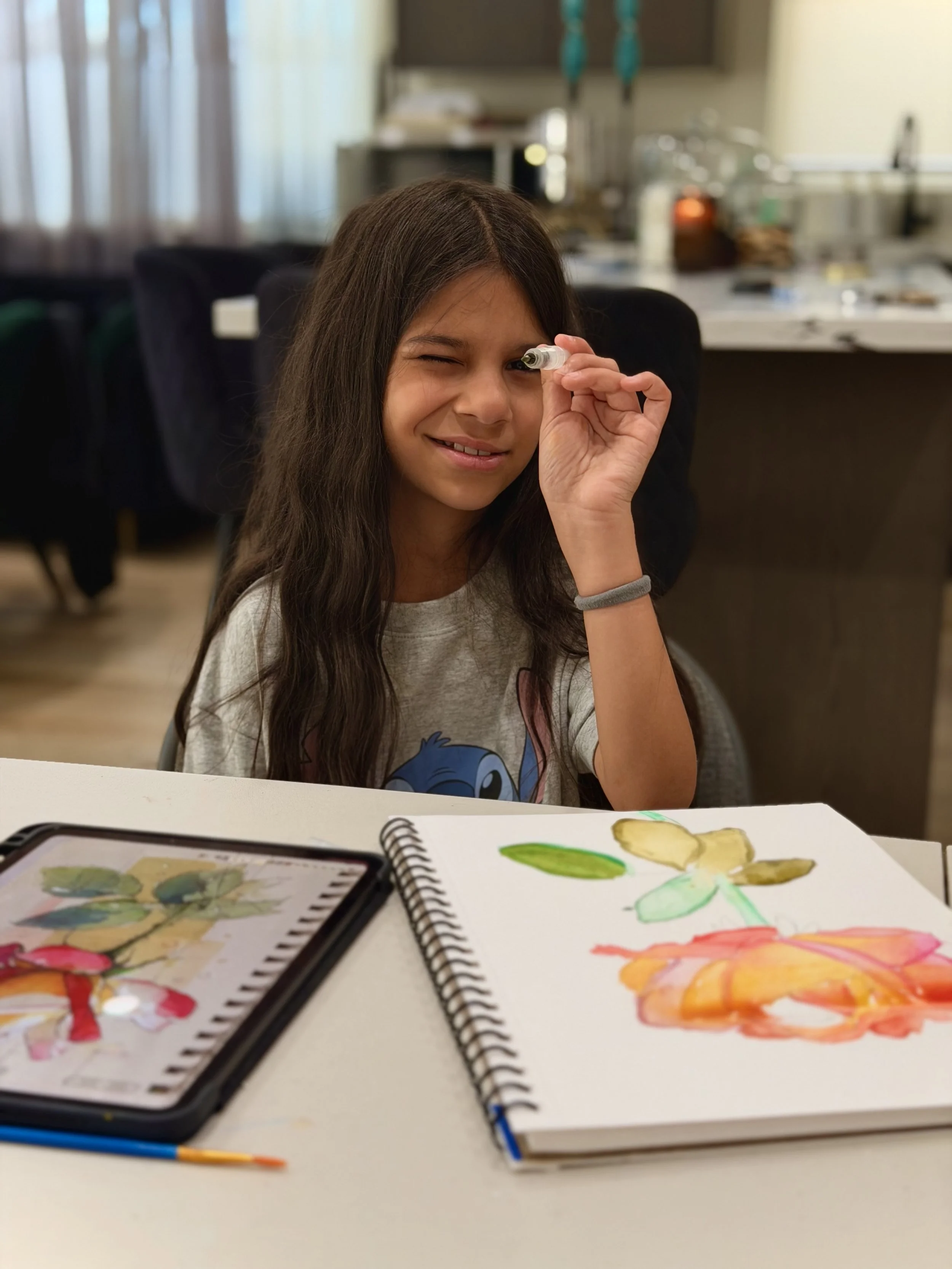 A young girl with long brown hair is winking and holding a small waterbrush near her eye. She is sitting at a table with sketchbooks, watercolor paints, and a paintbrush, with a background of a kitchen or dining area.
