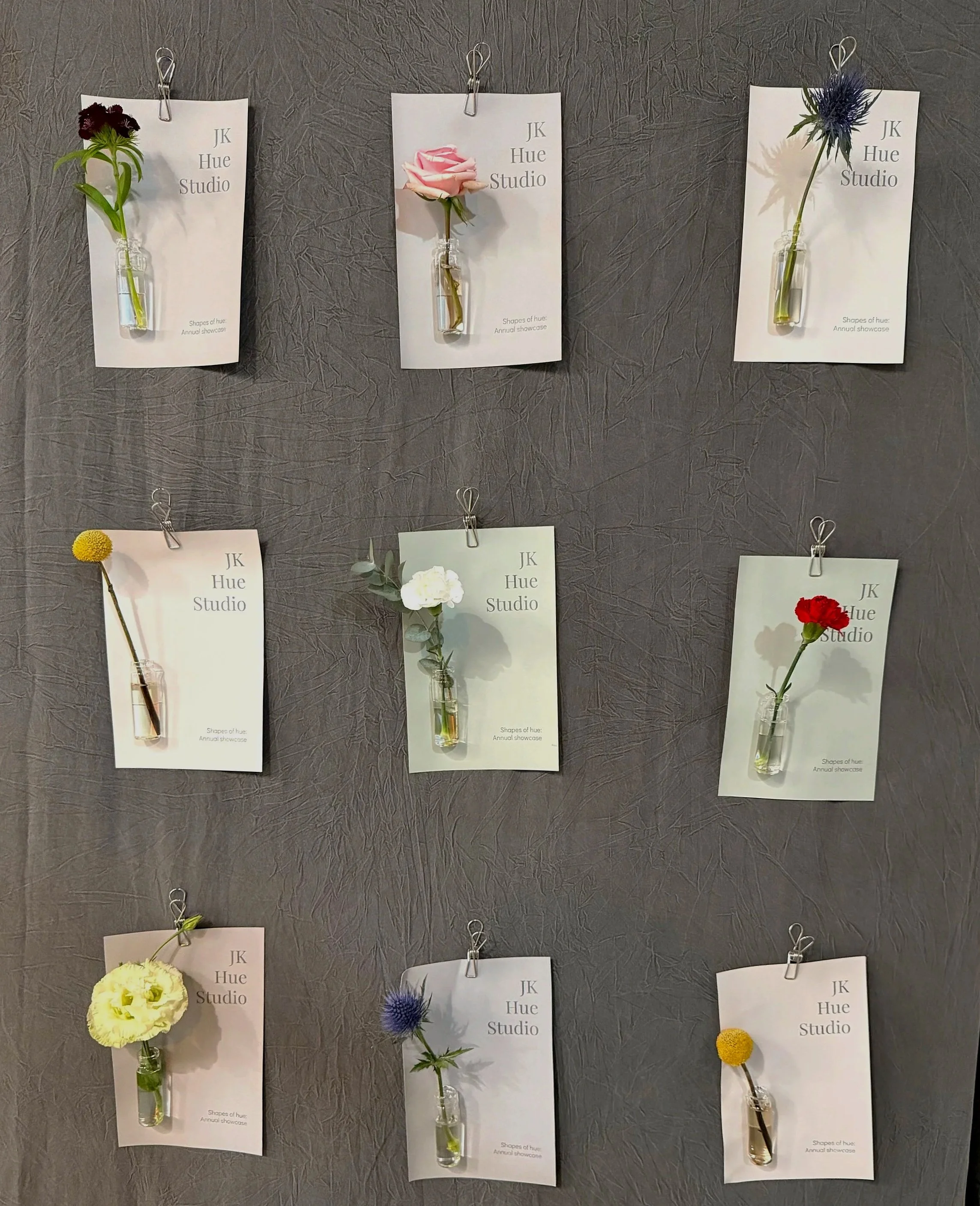 Display of six flowers in small glass vases attached to white cards with paper clips, arranged on a gray textured wall. The flowers include a dark red flower, a pink rose, a blue thistle, a yellow billy button, a white carnation, and a red carnation, each on a separate card labeled 'JK Hue Studio' with the text 'Shapes of hue: Annual showcase.'
