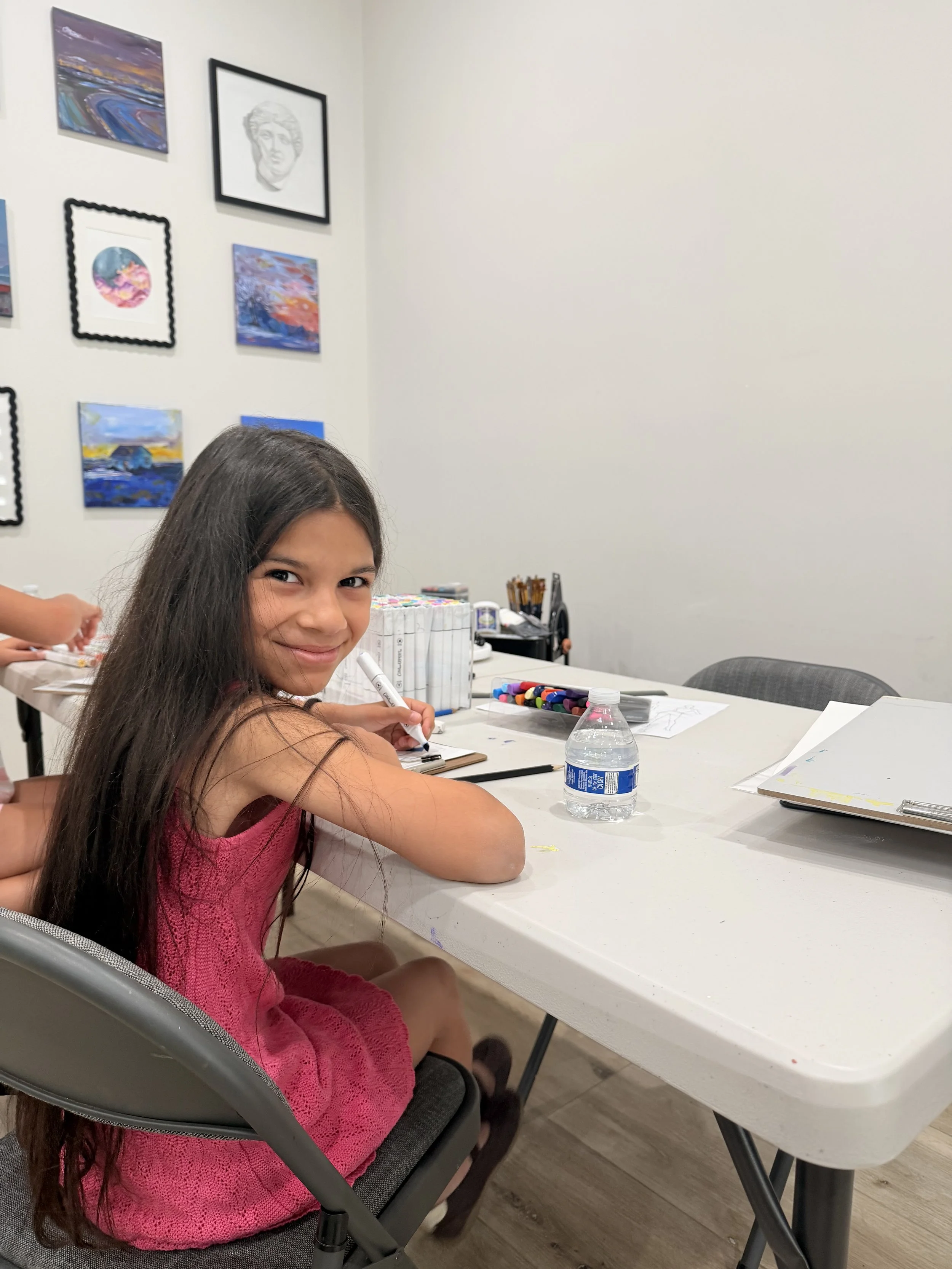 A young girl with long dark hair smiling while sitting at a white table, holding a marker. The table has art supplies, including markers and paper. Behind her is a white wall with various colorful paintings and drawings hanging.