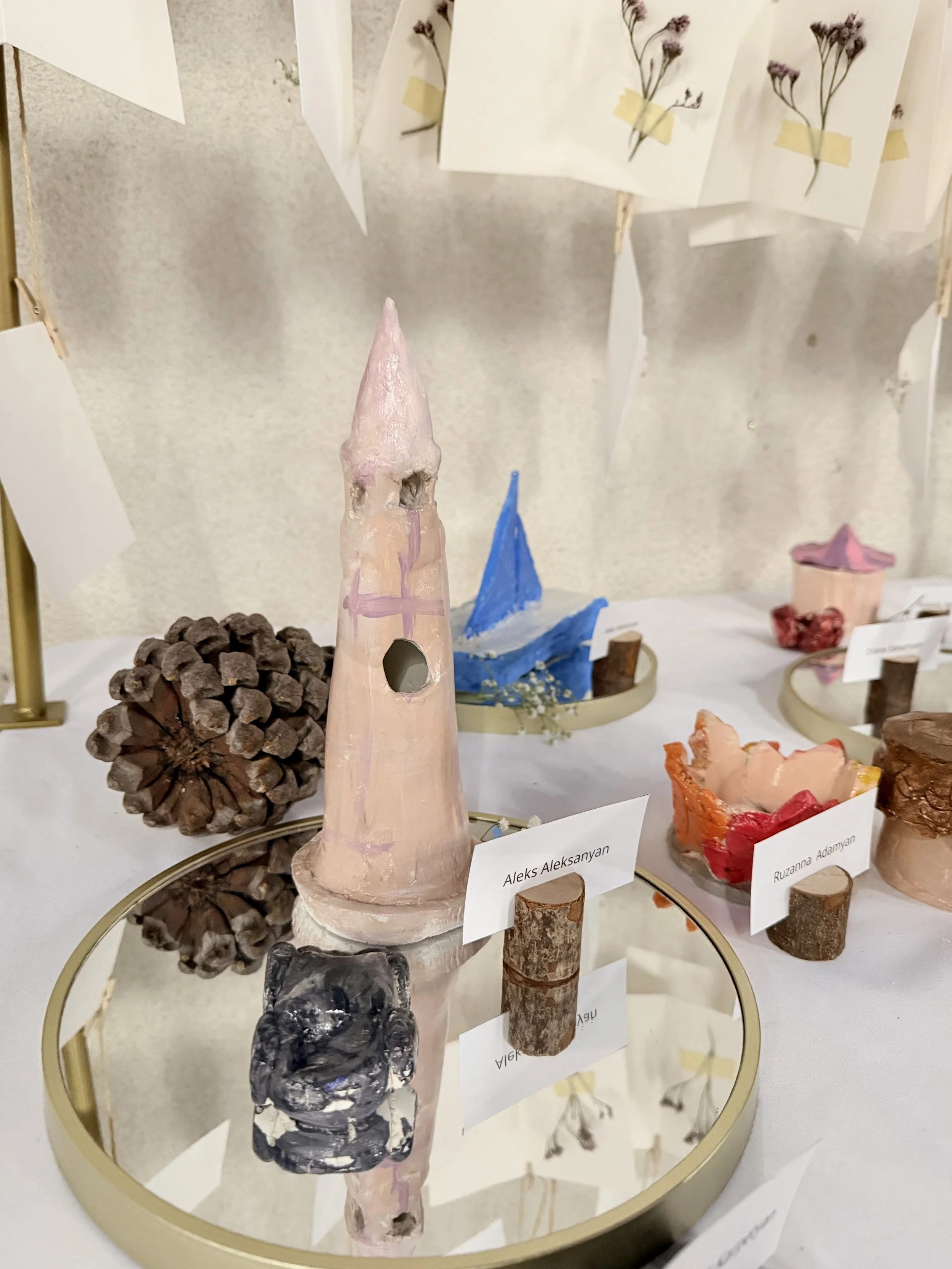 Various handmade ceramic sculptures resembling castles, spaceships, and abstract formations, displayed on a circular mirror surface with small labels in front of each piece.
