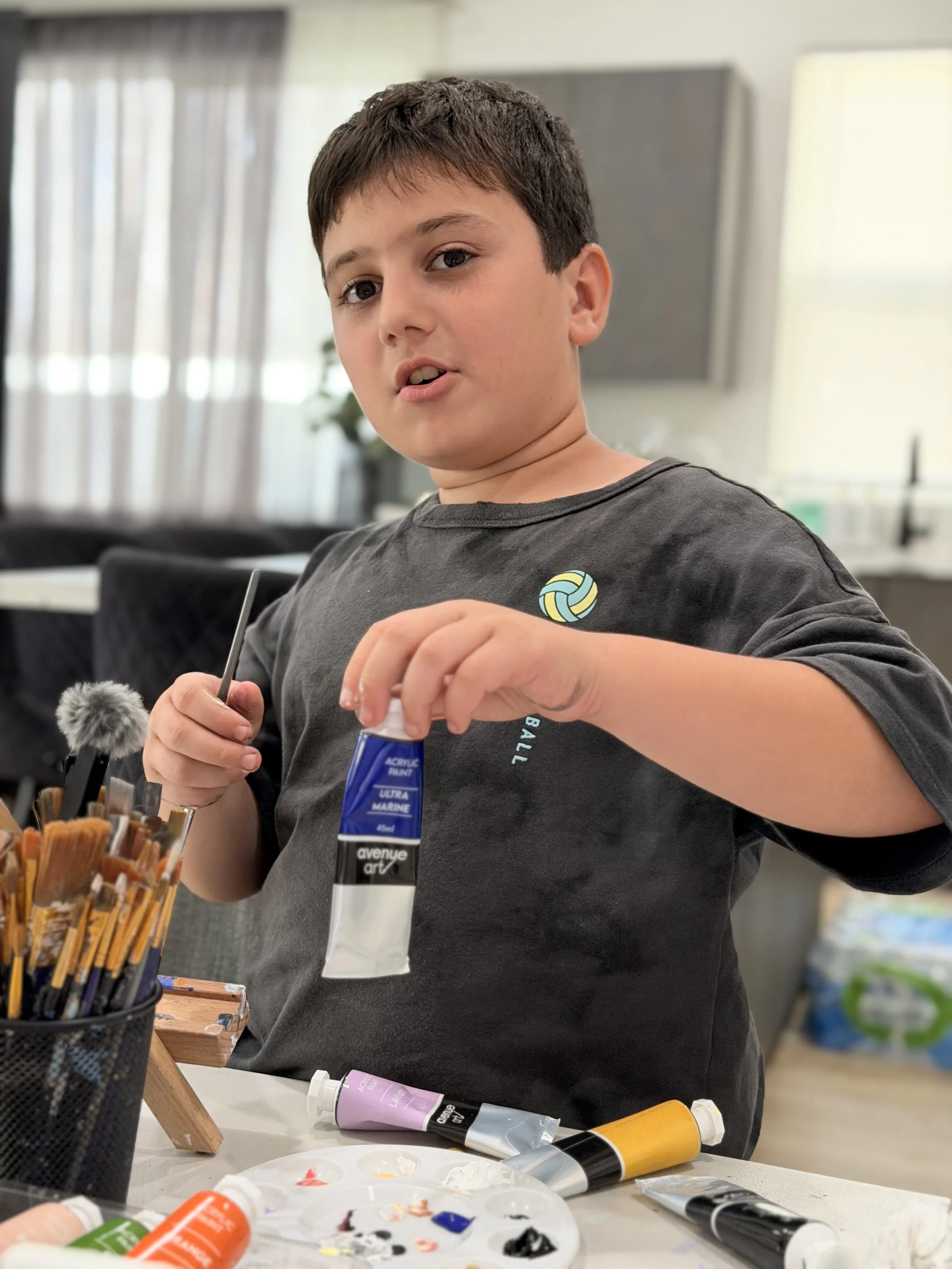 A young boy with dark hair and a gray T-shirt is sitting at a table, holding a tube of acrylic paint. Various art supplies, including paint tubes, brushes in a holder, and a palette with mixed paint colors, are on the table. The background shows a br