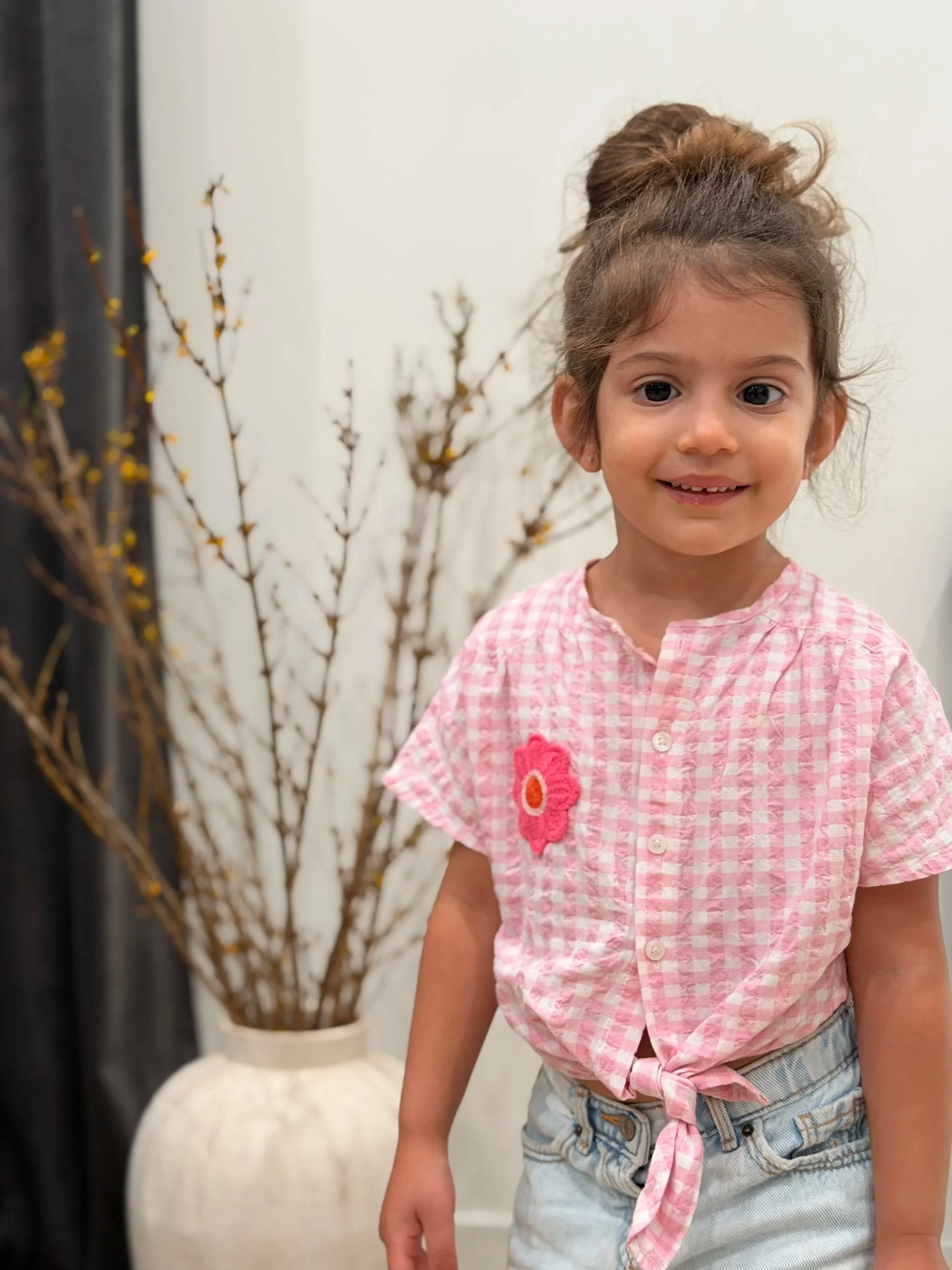 Close-up of a smiling young girl with brown hair tied in a bun, wearing a pink and white checkered shirt with a pink flower patch, and denim shorts, standing indoors in front of a decorative branch arrangement.