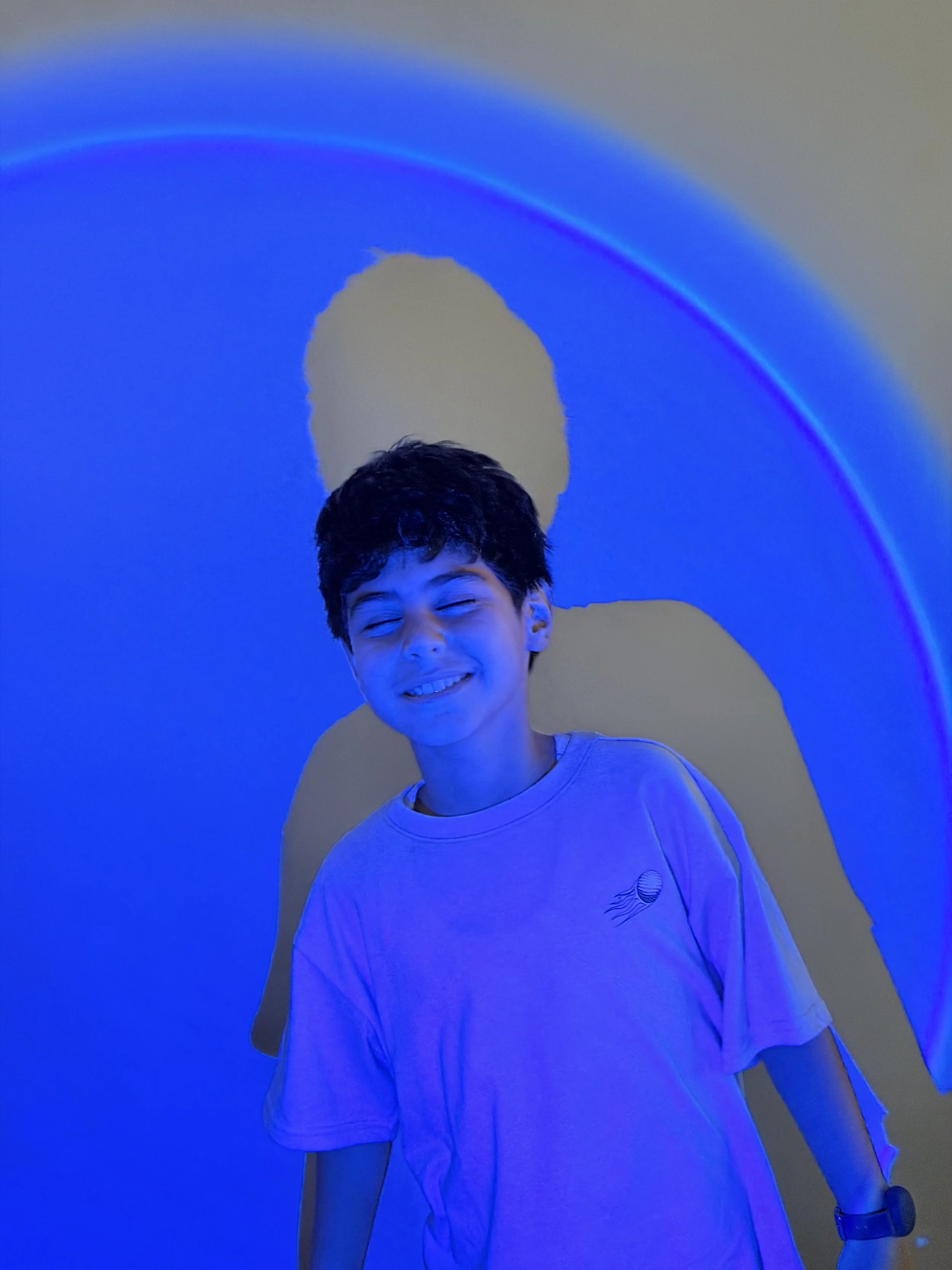A smiling boy with dark hair is illuminated by blue and purple light, casting shadows on the wall behind him.