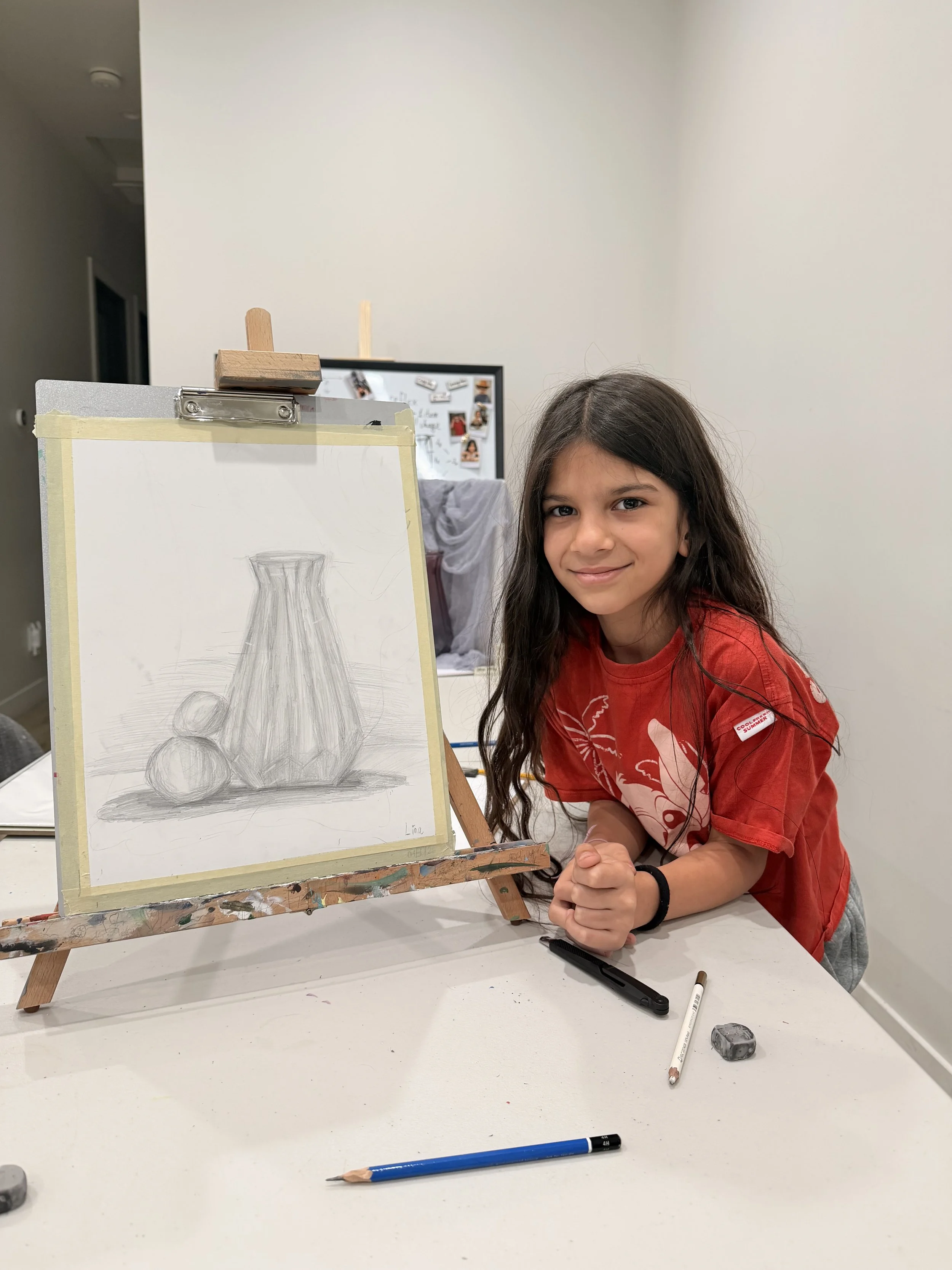 A young girl with long dark hair and a red shirt is smiling and leaning on a table next to a drawing of a still life with a vase and three round objects on a white sheet of paper on an easel. Art supplies such as a pencil, marker, and eraser are on t