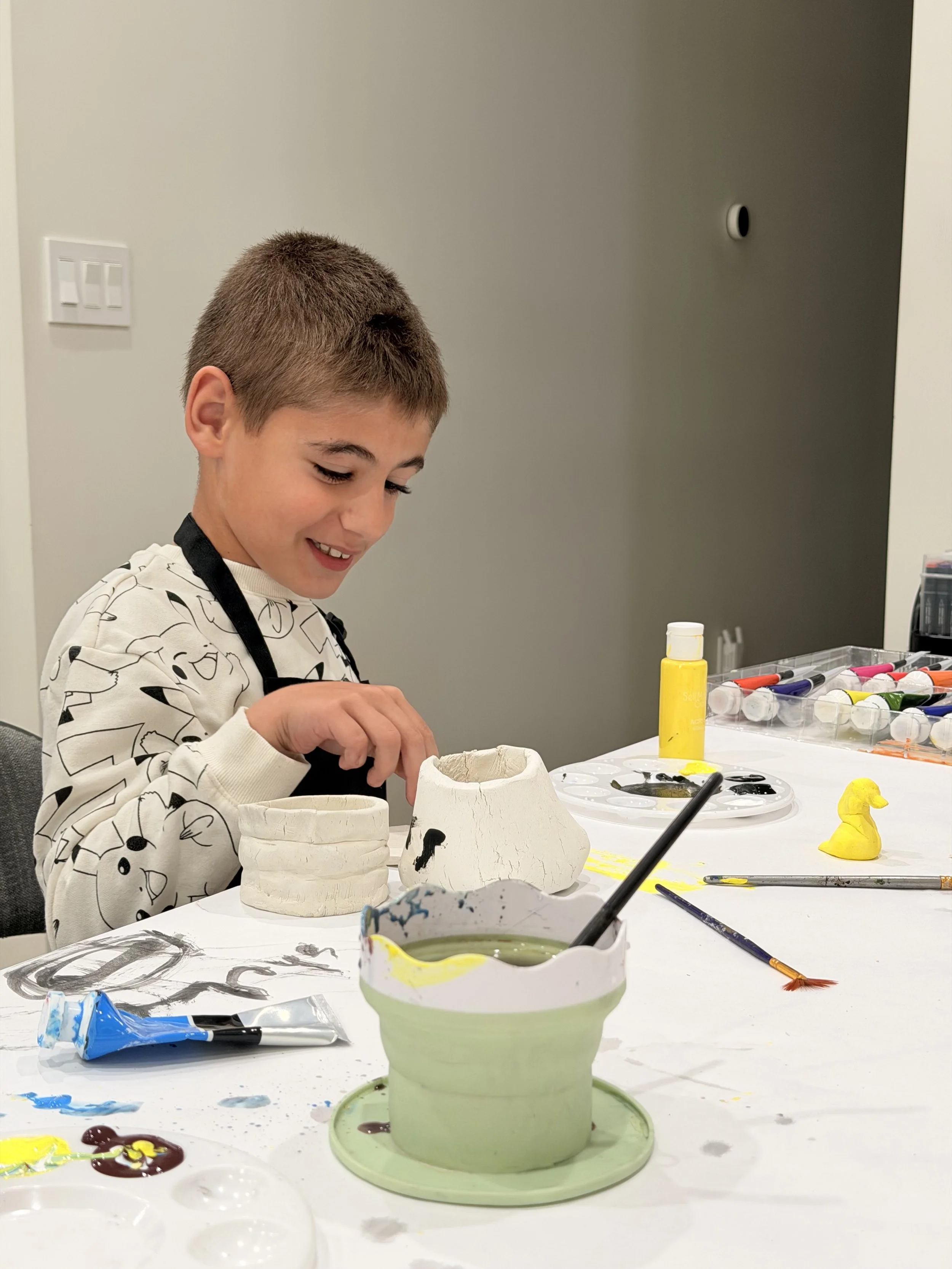 A young boy with short brown hair and a smile is painting ceramic items at a table. The table is covered with paint supplies, including containers of paint, brushes, and painted items such as a yellow duck. The boy is wearing a white shirt with black