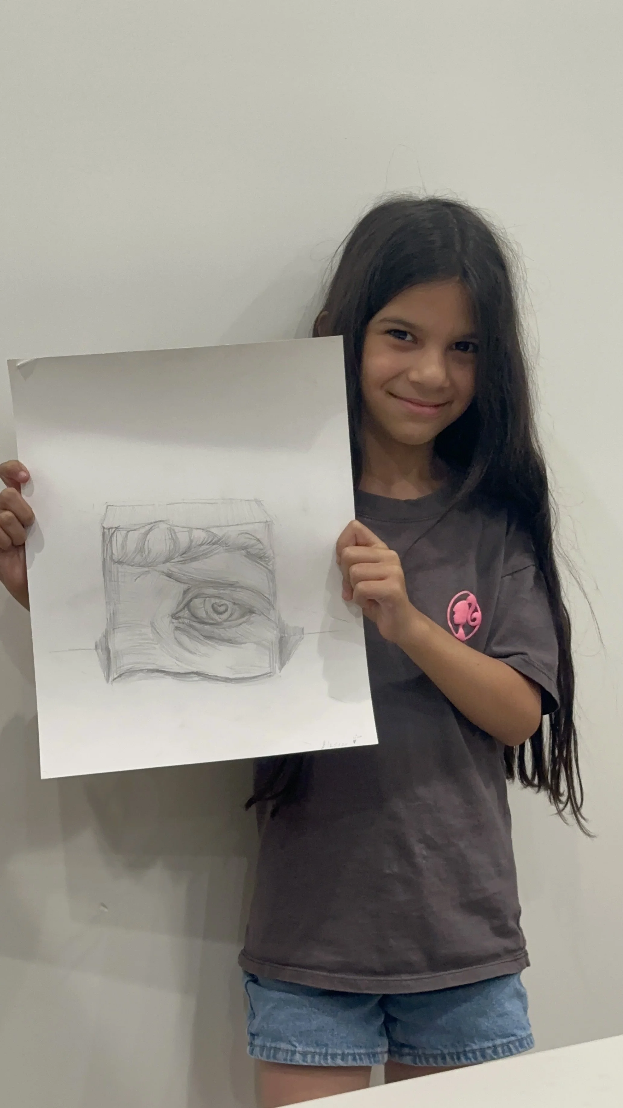 Young girl with long dark hair holding a sketch of an eye on paper, smiling, wearing a gray T-shirt and denim shorts, standing against a plain white wall.