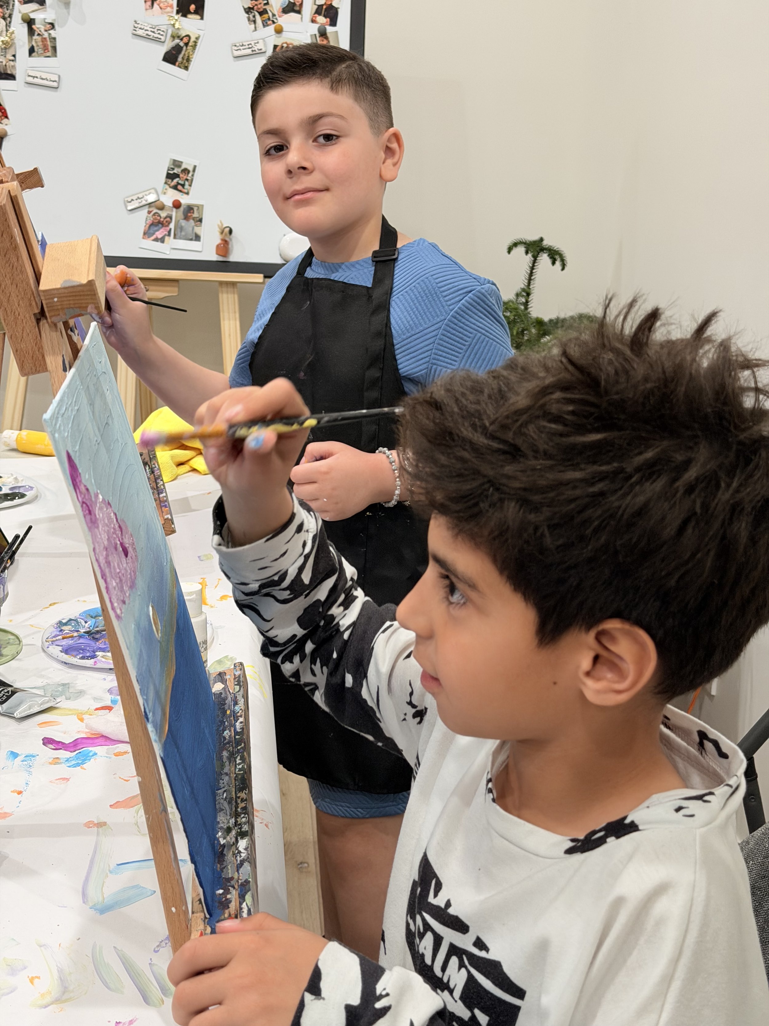 Two boys painting on canvas at a table. The boy in the background has short dark hair, a blue shirt, and a black apron. The boy in the foreground has dark hair, a striped shirt, and is holding a paintbrush, painting on the canvas.
