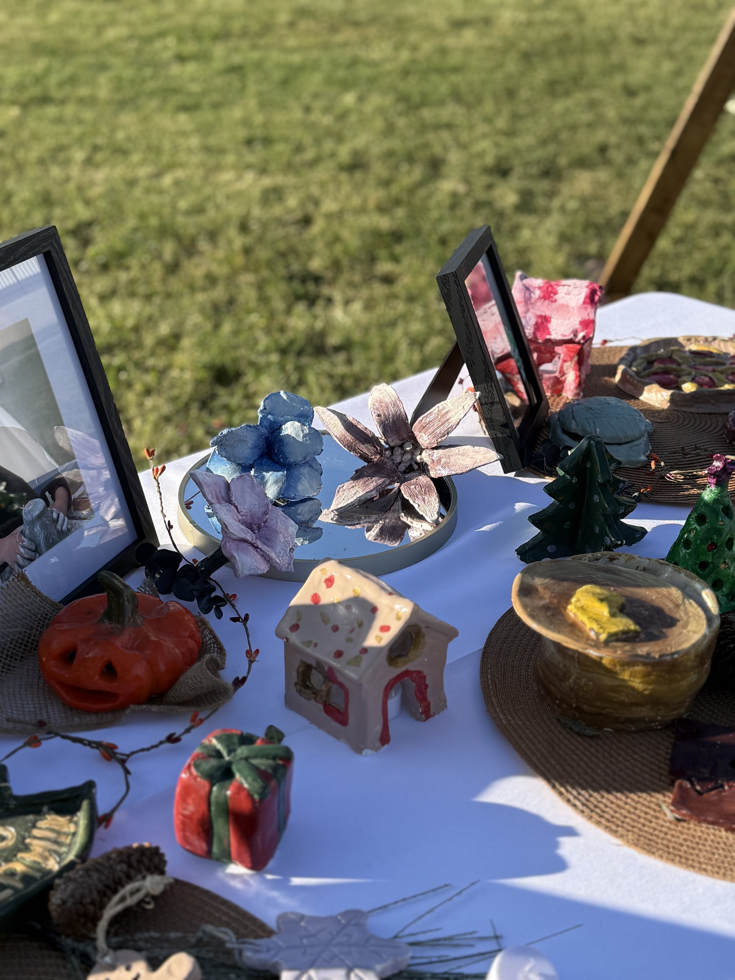 Display of handcrafted ceramic and glass art pieces on a table outdoors, including flowers, animals, and decorative objects.