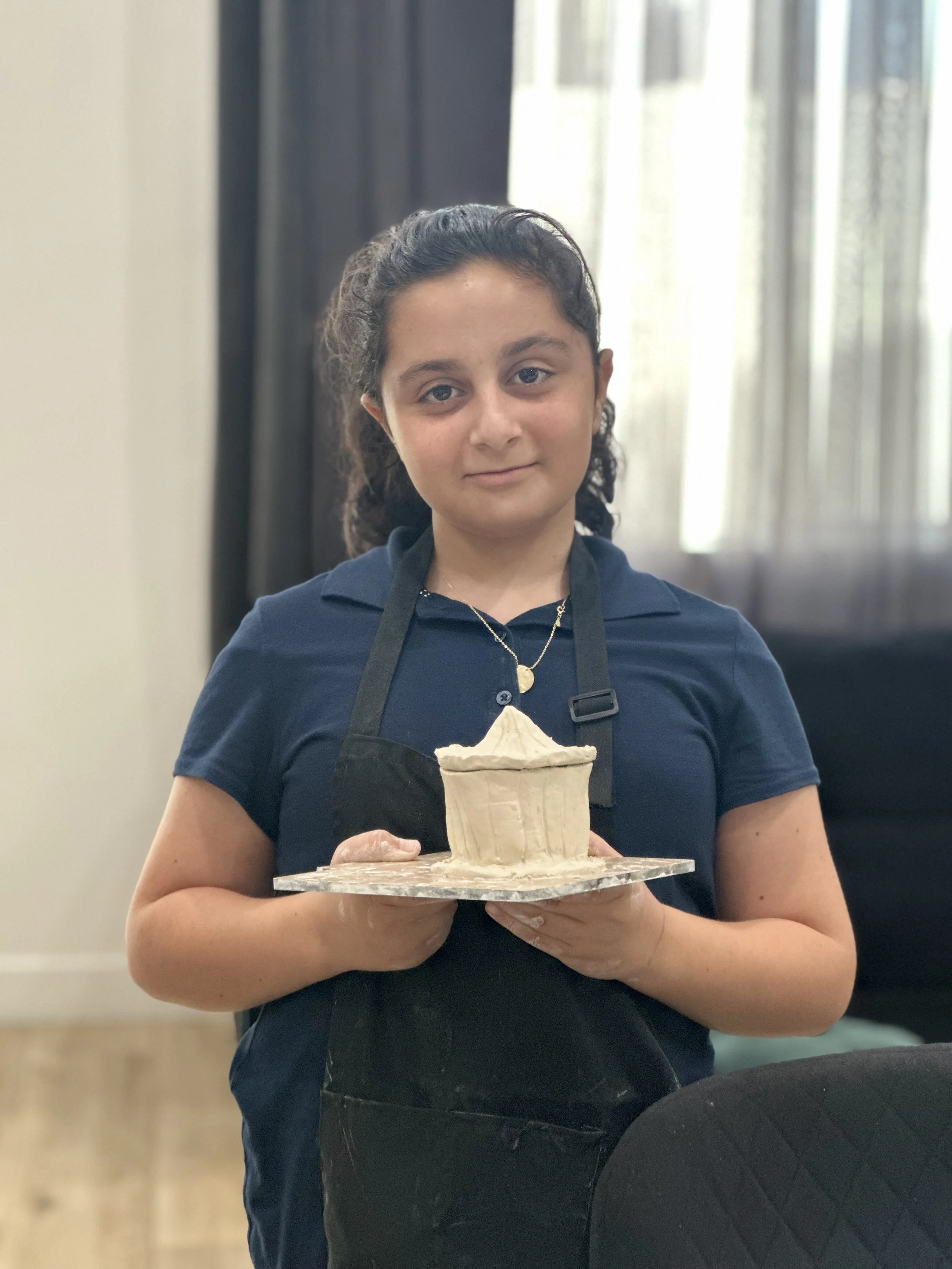 A girl with curly hair and a dark blue shirt holding a large ice cream cone covered in white frosting, standing indoors near a window with curtains.