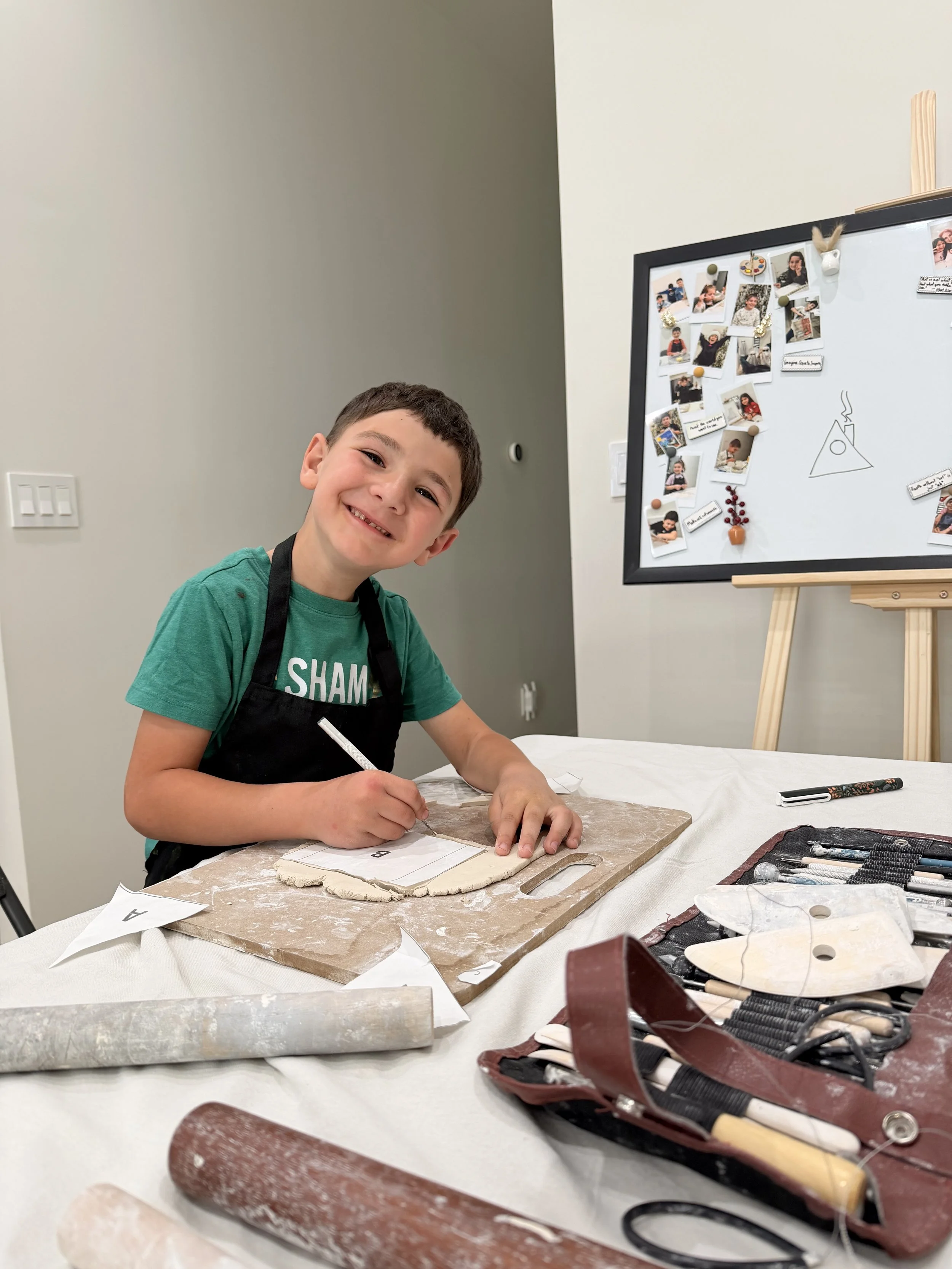 A young boy smiling at the camera while working on a pottery project at a table with various pottery tools and materials.