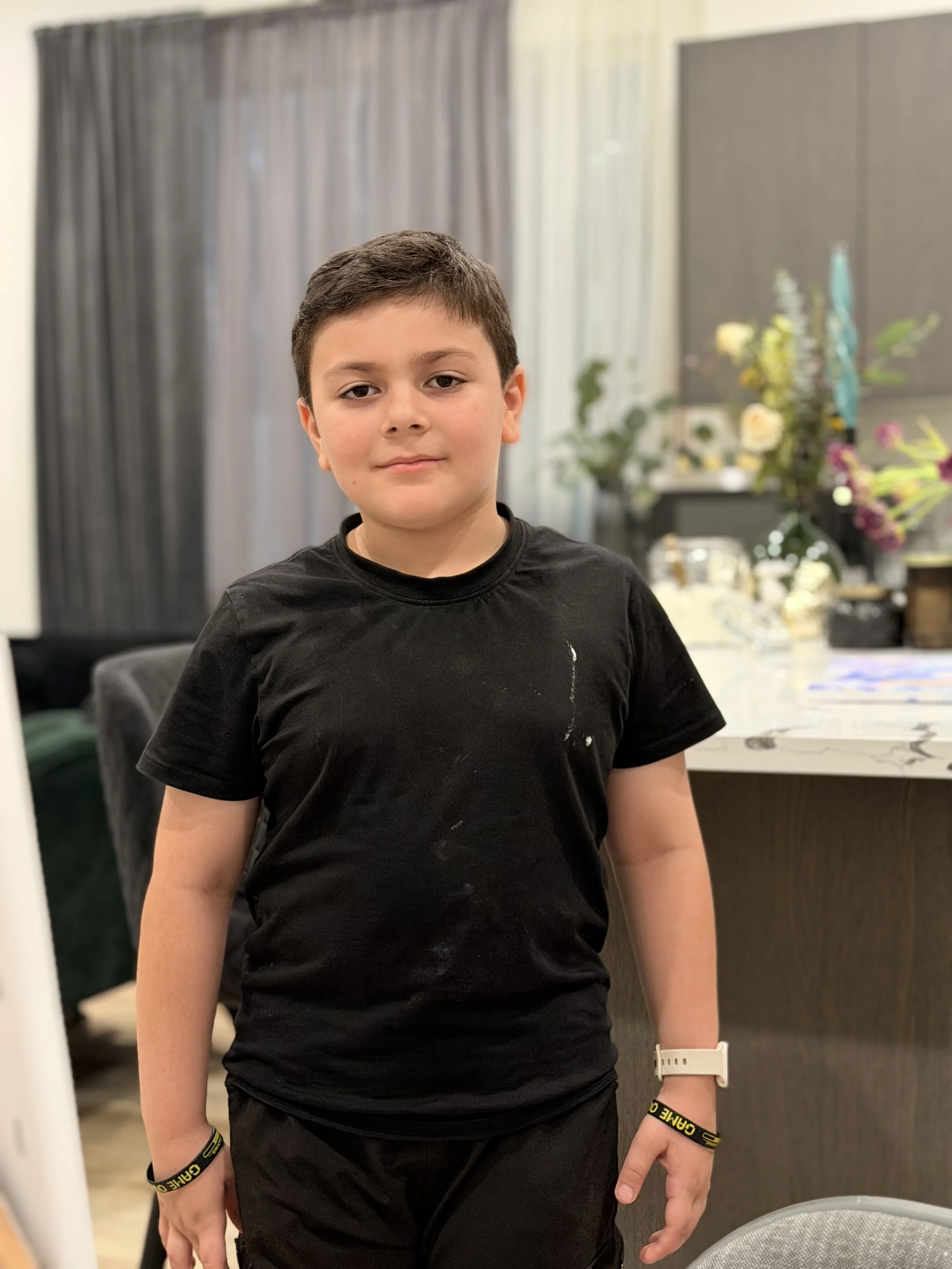 A young boy with short dark hair, wearing a black t-shirt and black shorts, standing indoors with a neutral expression.
