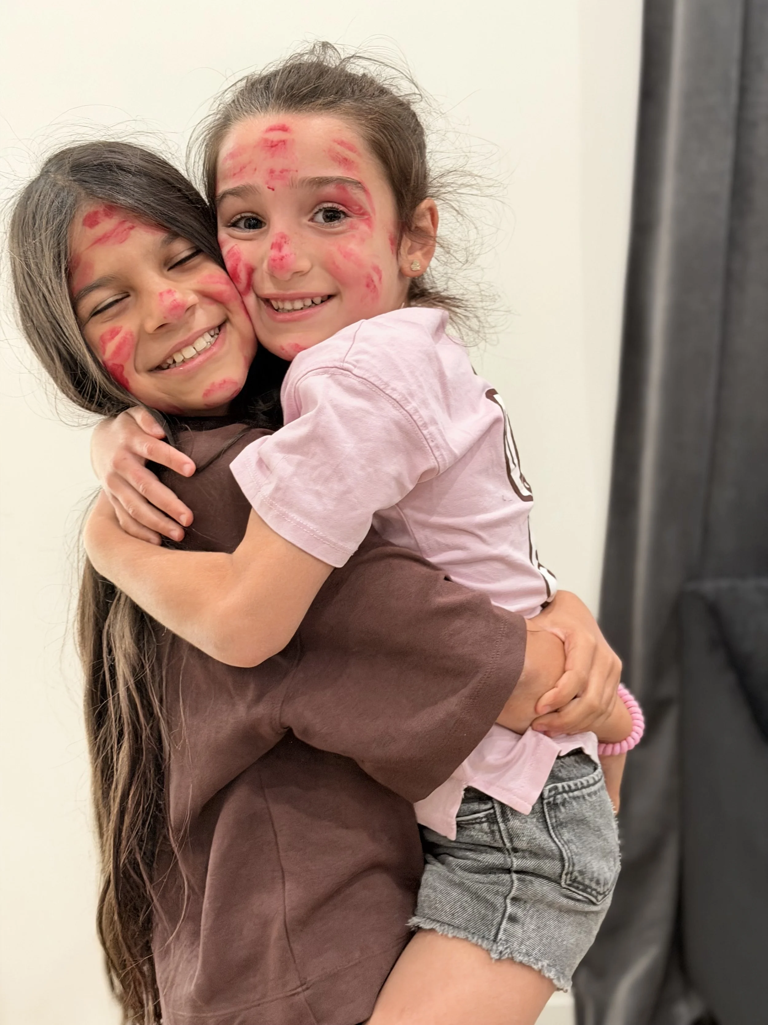 Two young girls hugging each other with red lipstick marks on their faces, smiling, in front of a plain wall.