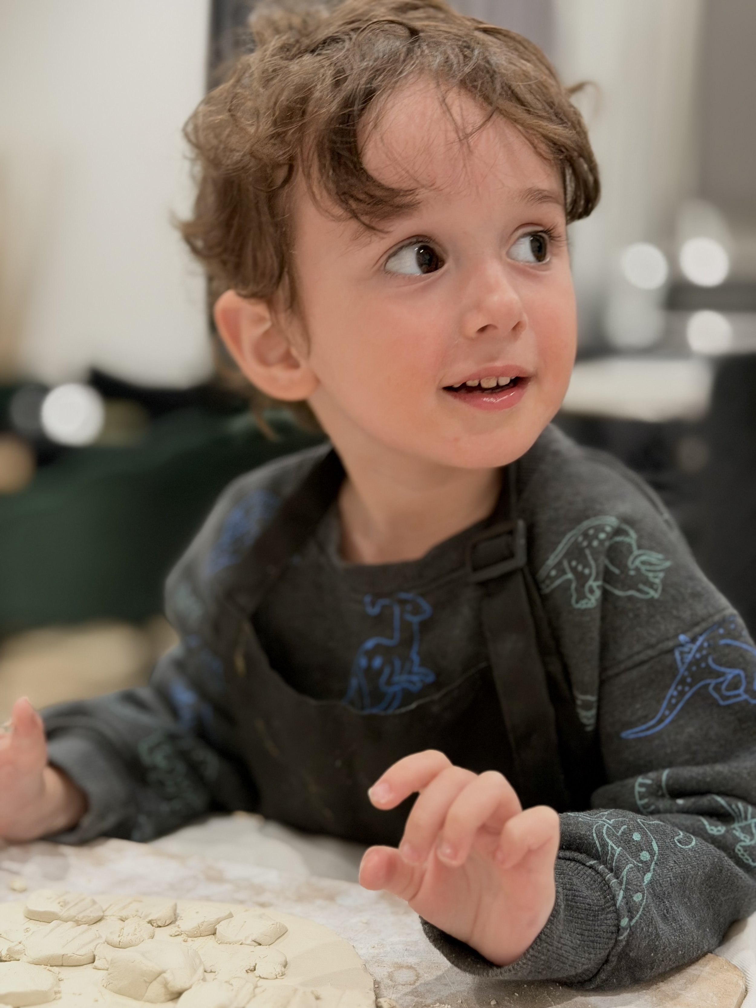 A young boy with brown curly hair and light skin is lying on a table, looking to his right with a curious expression. He is wearing a dark gray hoodie with blue patterns of dinosaurs. There is white dough or clay and small pieces on the table in fron