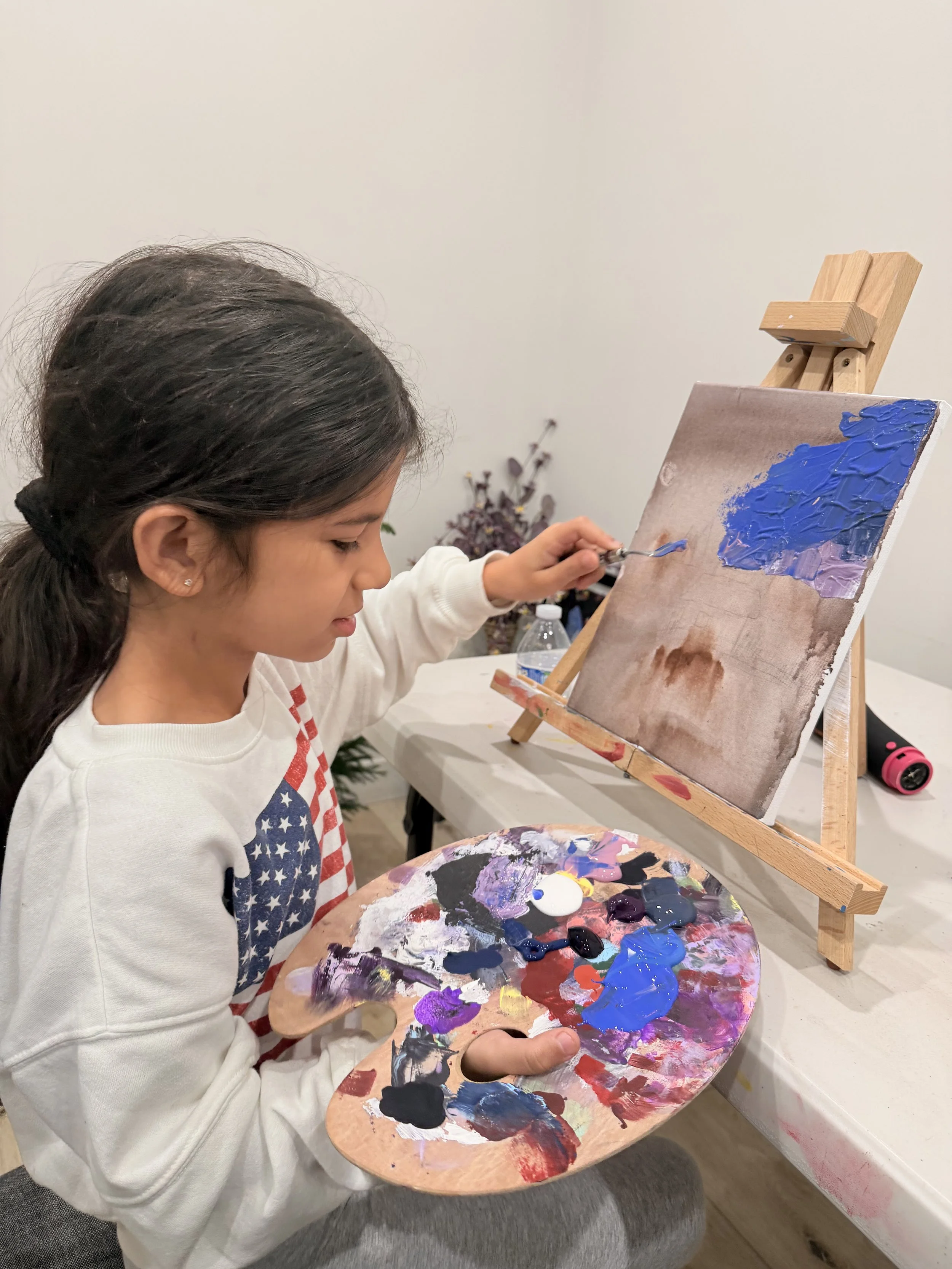 A young girl with dark hair tied back, wearing a white sweatshirt with an American flag design, is painting on a canvas. She holds a wooden palette with various colors of paint and is applying purple to her artwork. The canvas is on an easel, and the