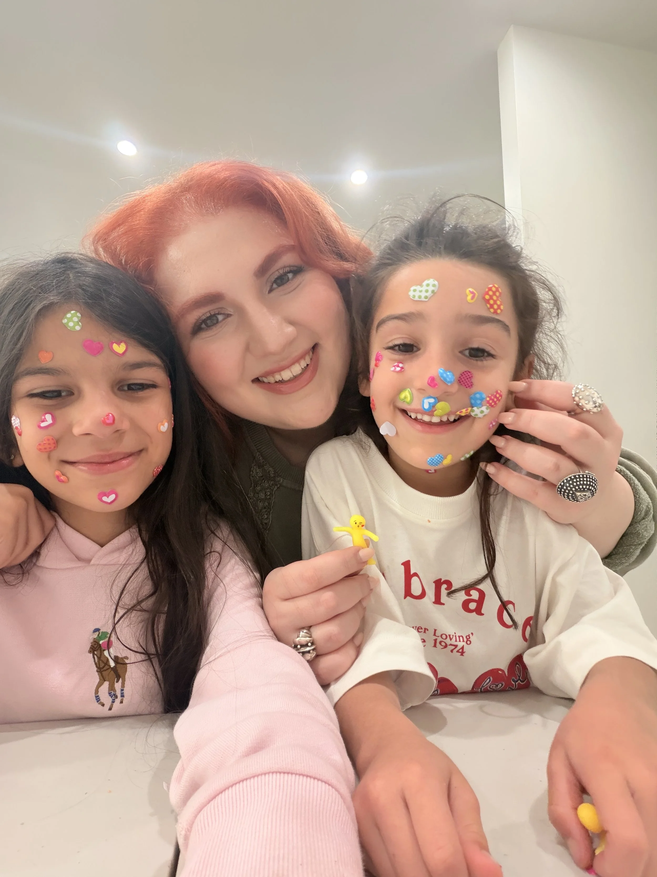 Three women smiling closely together, with two children also smiling, all with colorful heart and dot stickers on their faces, in an indoor setting with white walls and ceiling lights.