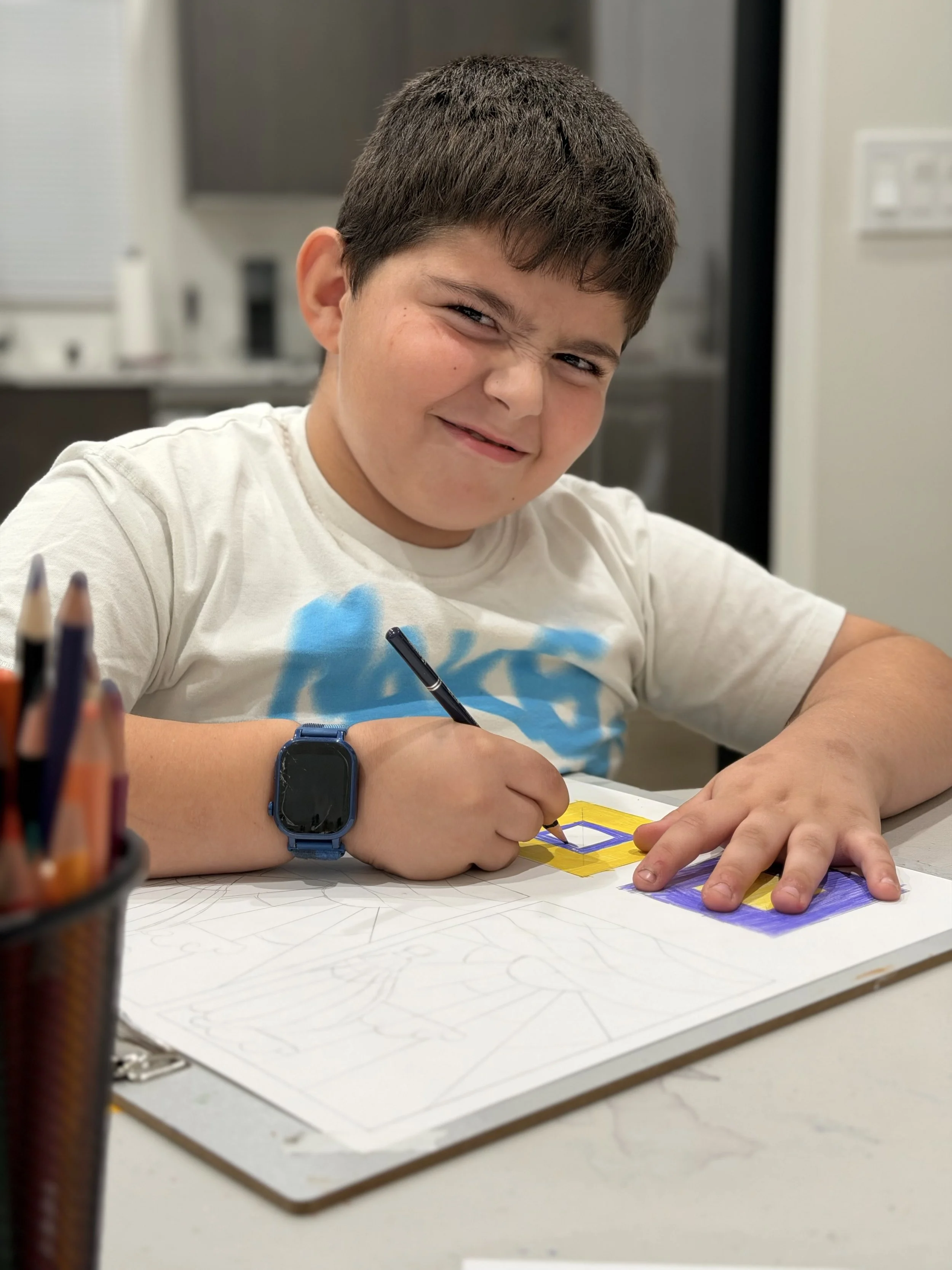 A young boy with short brown hair and a blue smartwatch on his left wrist is smiling mischievously while coloring geometric shapes on a large sheet of paper with colored markers. The boy is wearing a white t-shirt with blue and black designs, and is sitting at a white table with a container of colored markers in the foreground. The background is a kitchen.