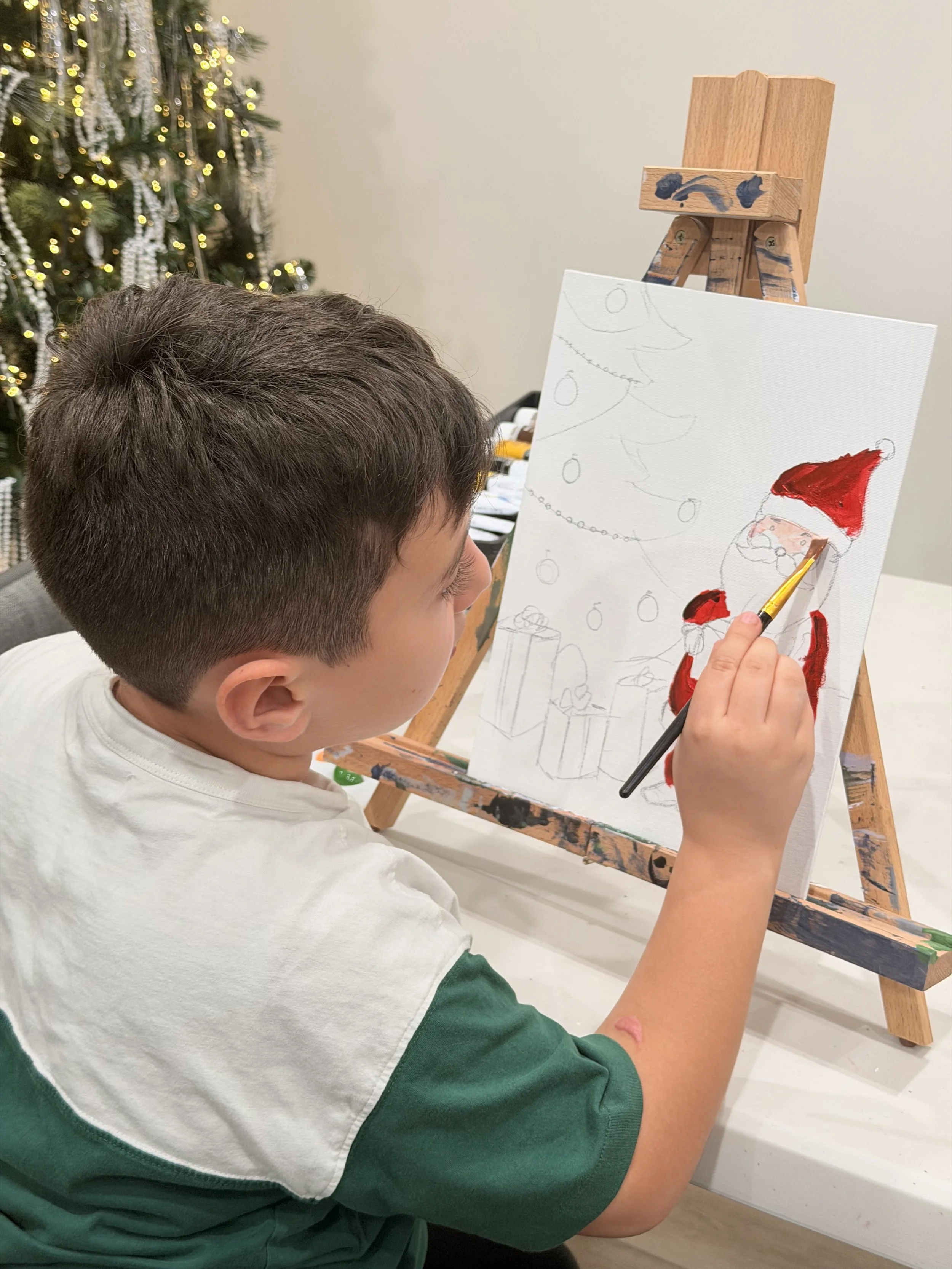 A boy in a green and white shirt is painting a Santa Claus on a canvas, with a Christmas tree decorated with lights and ornaments in the background.