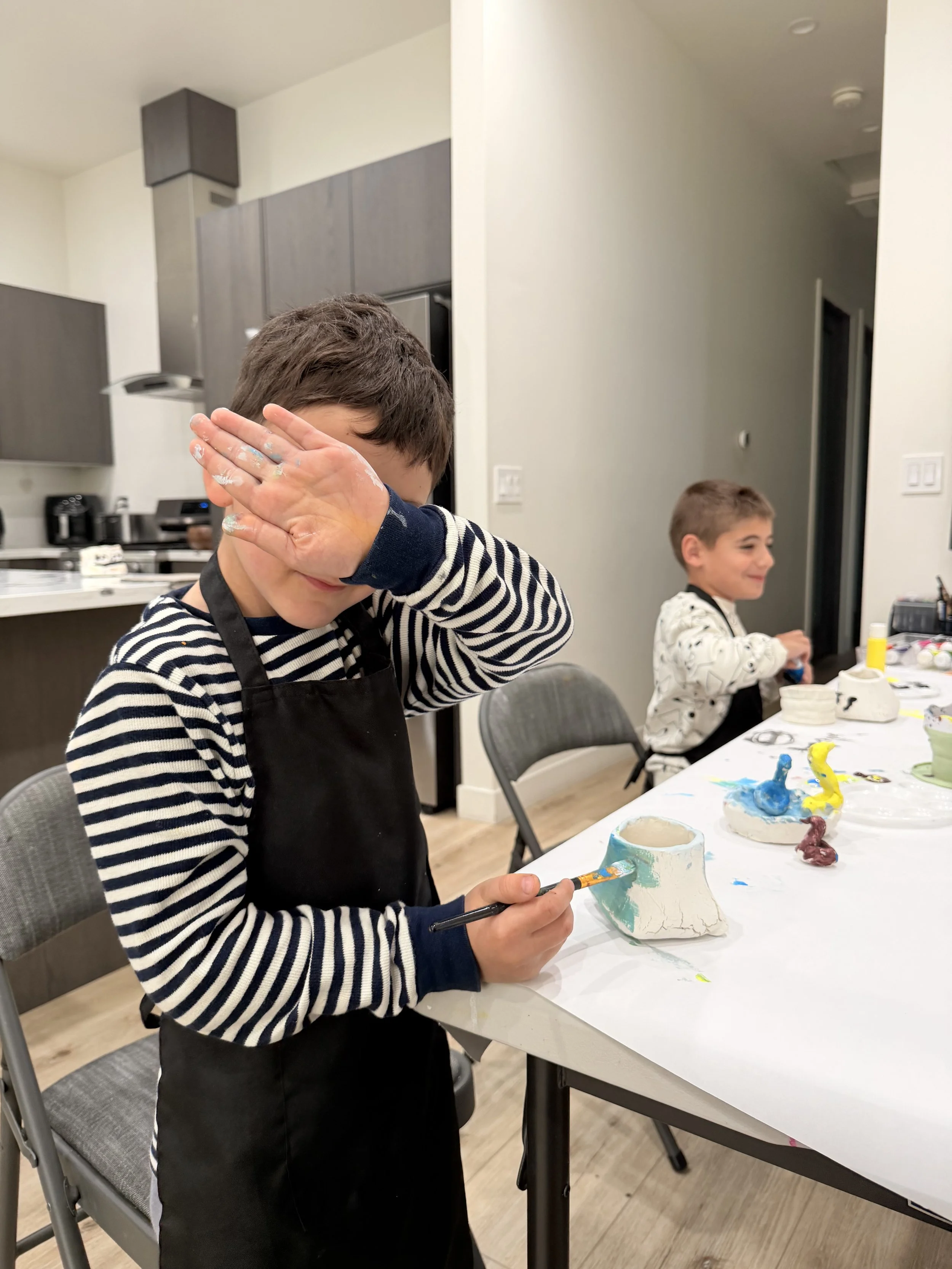 Two boys are participating in a ceramics painting activity at a table covered with paint and clay sculptures. One boy, in a striped shirt and black apron, is covering his face with one hand while holding a paintbrush and a ceramic piece. The other bo