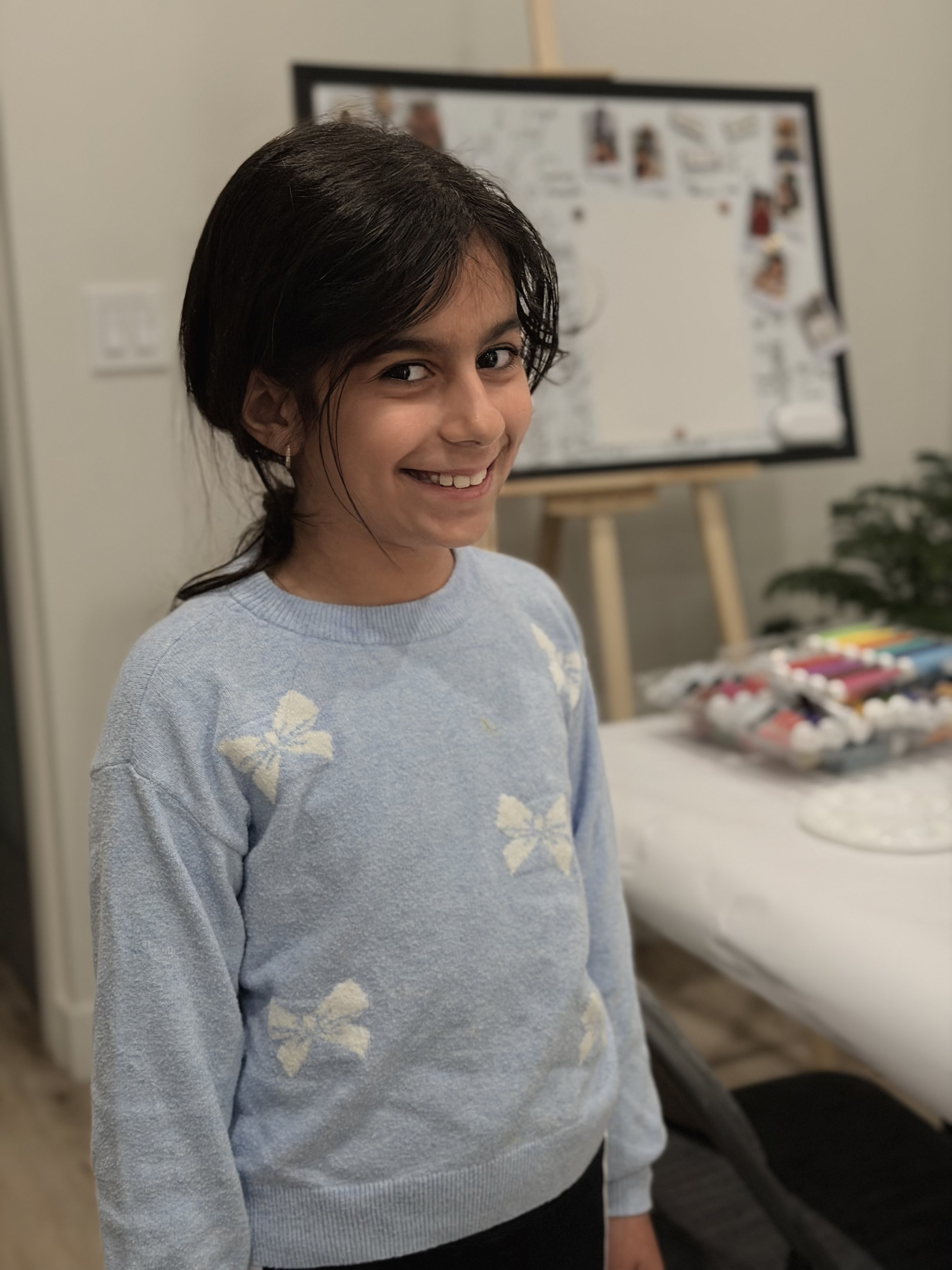 A young girl with dark hair, wearing a light blue sweater with white floral patterns, smiling at the camera in an indoor setting with a blurred background including a whiteboard or display with photos and colorful objects.