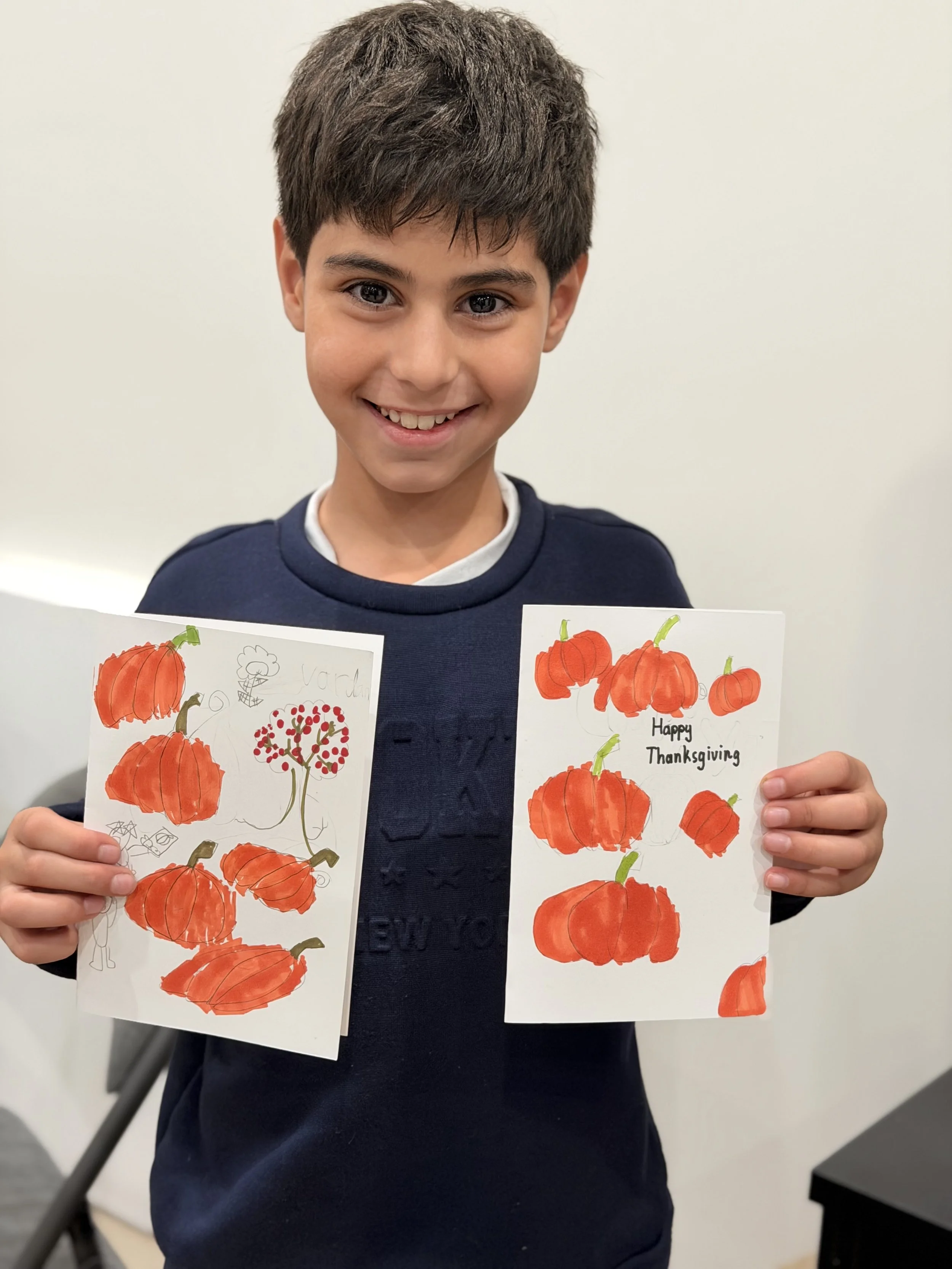 A young boy smiling and holding up two handmade Thanksgiving cards with orange pumpkins and some red accents.