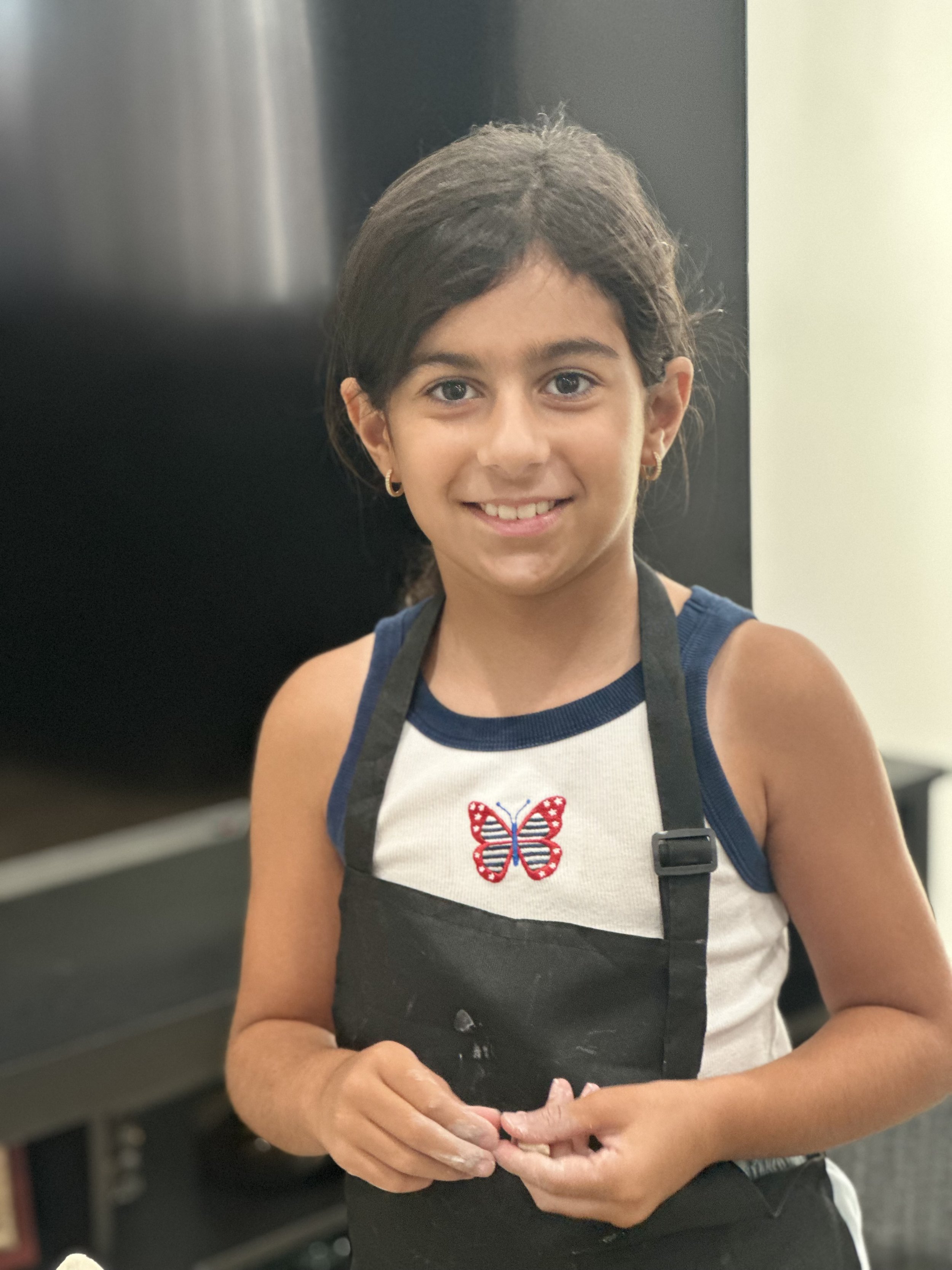 A young girl with dark hair, wearing a white shirt with a red, white, and blue butterfly emblem, and a black apron, smiling and holding her hands together.