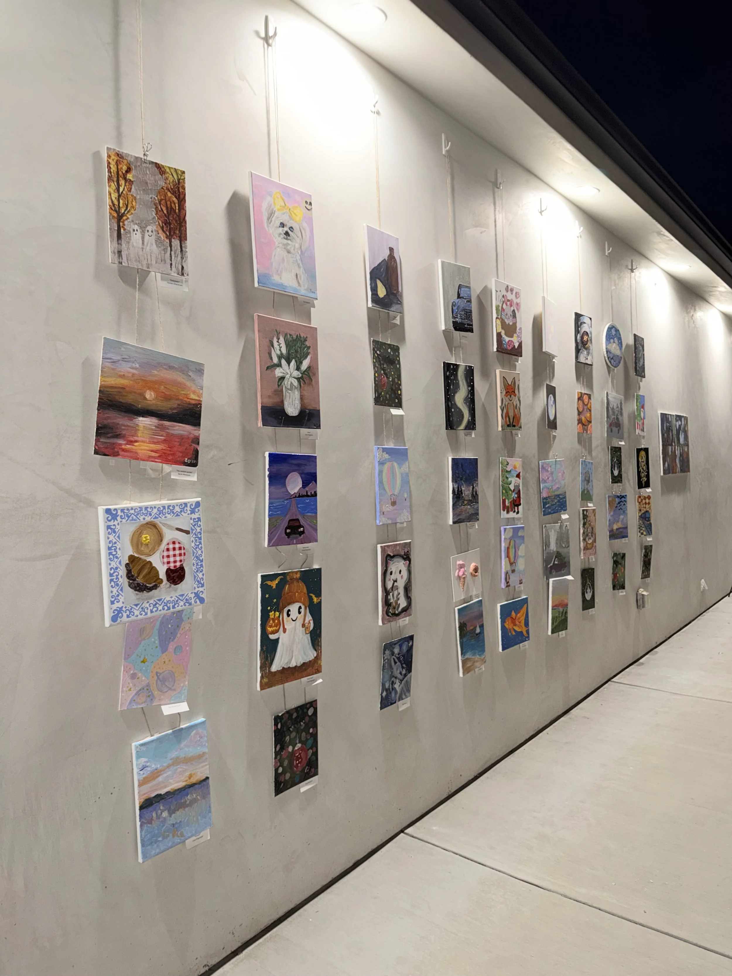 Wall displaying multiple small colorful paintings hung in a grid pattern on a gallery wall.
