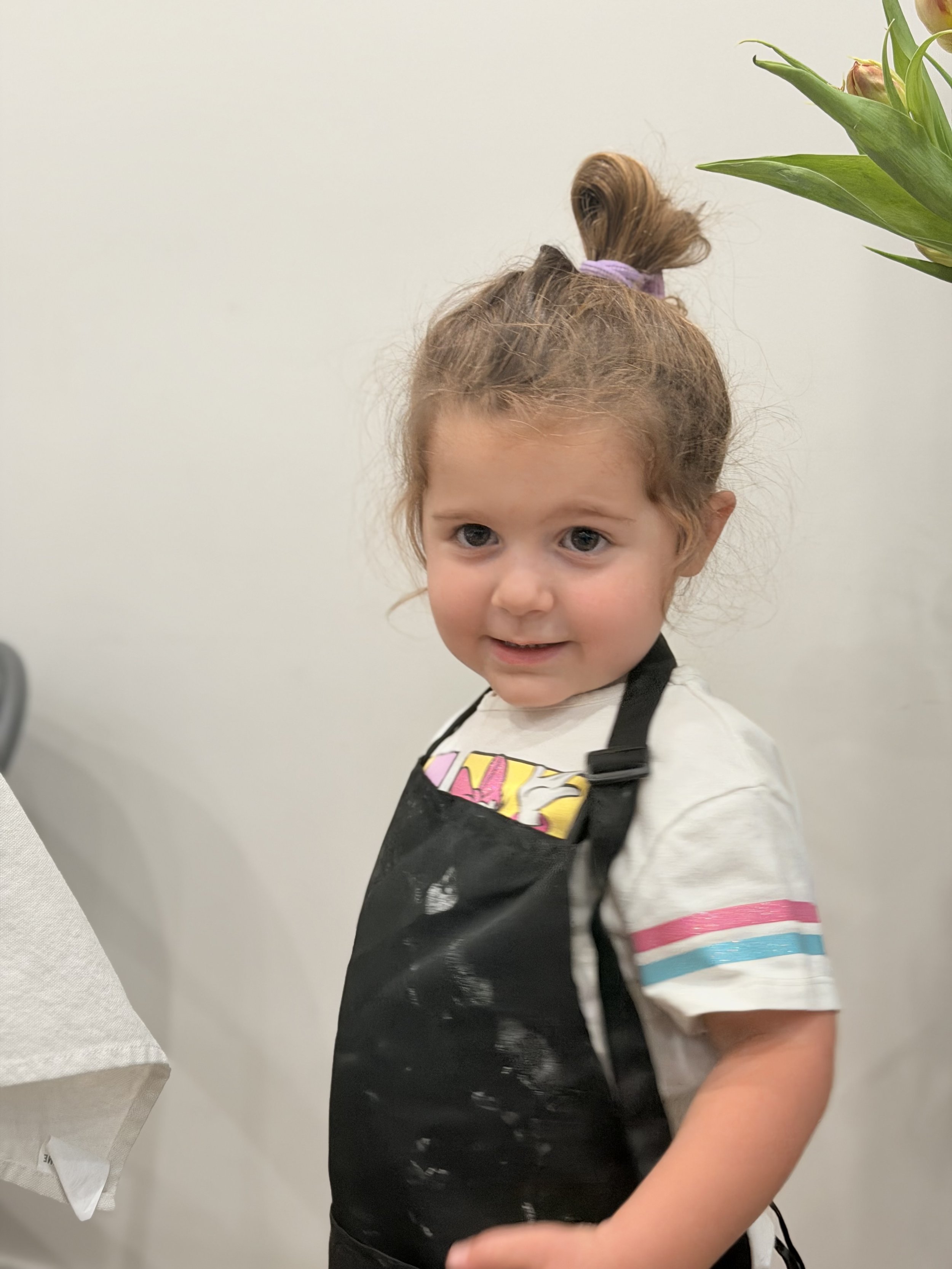 A young girl with curly light brown hair tied up with a purple hair tie, wearing a white t-shirt with colorful stripes on the sleeves, and a black apron or vest. She is smiling slightly, standing next to a white wall with a partial view of a potted p