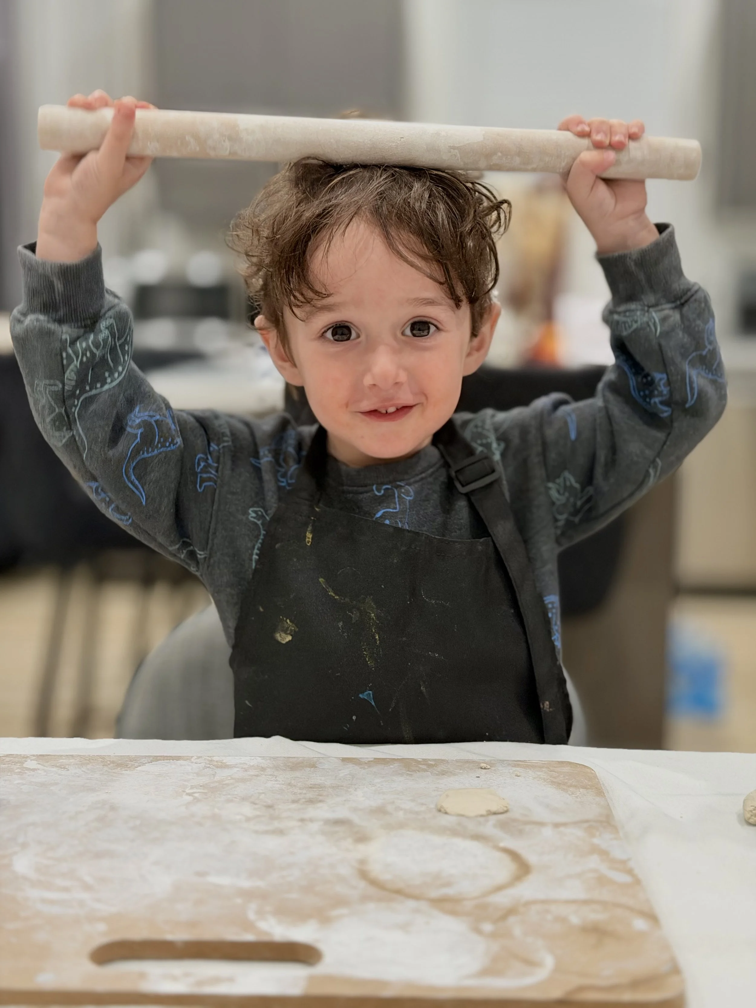 A young boy with curly brown hair, wearing a gray long-sleeve shirt and a black apron, is holding a rolling pin above his head in a kitchen. In front of him, a piece of dough sits on a floured surface.