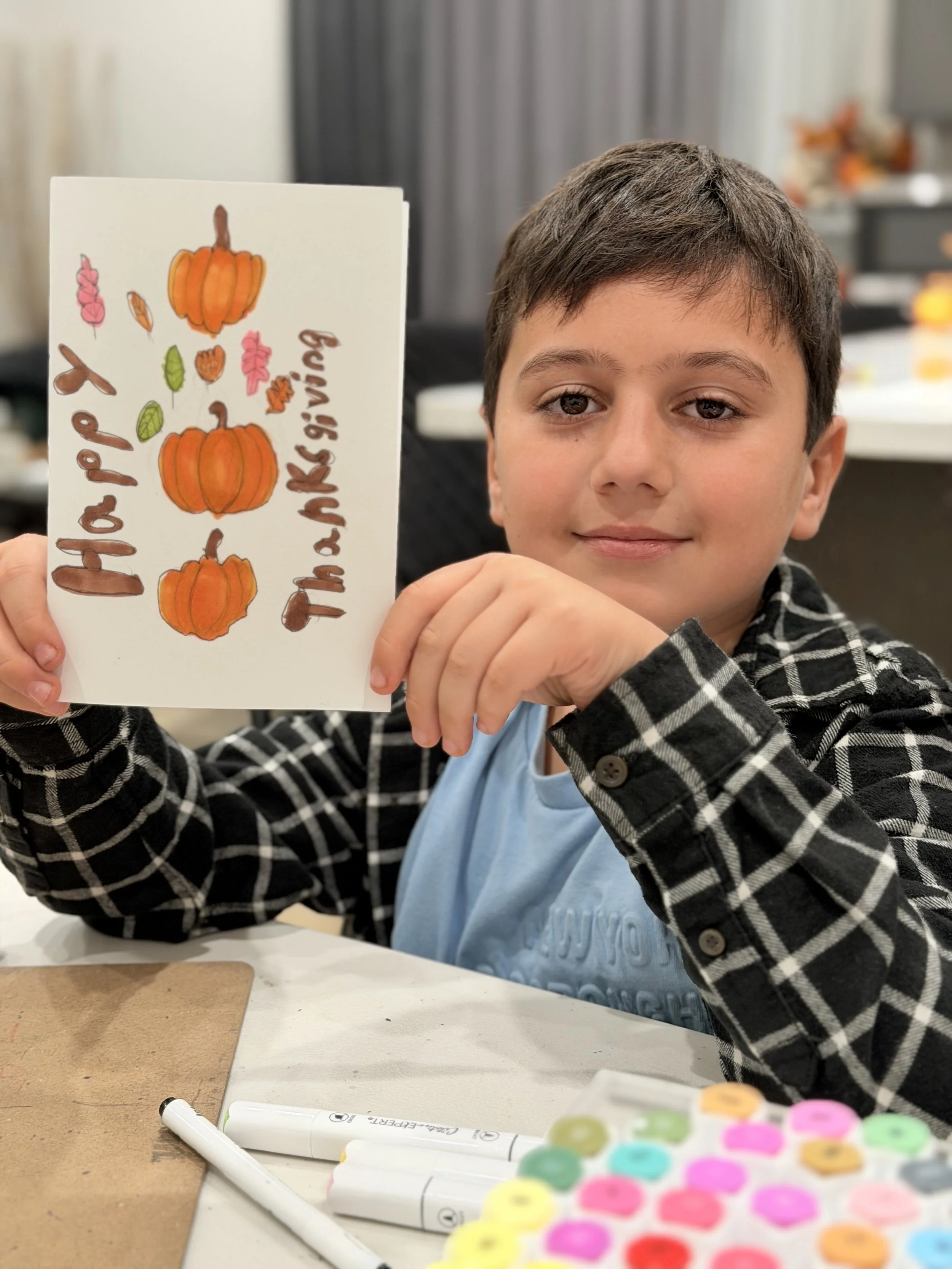 A young boy with brown hair and a checkered jacket holding a handmade card with pumpkins and fall-themed drawings, sitting at a table with markers and colorful stickers.
