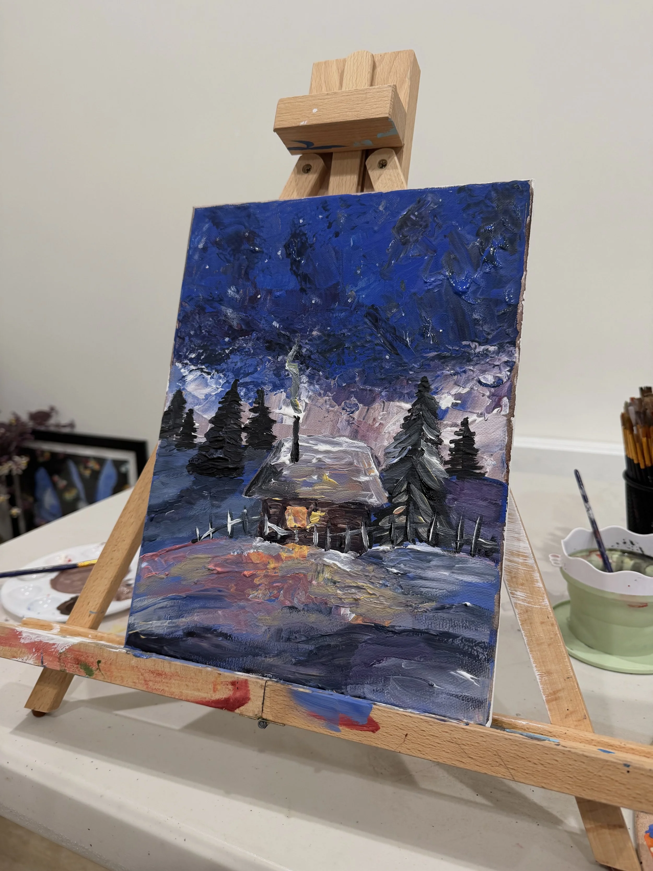 Painted landscape scene featuring a house with a lit window, tall pine trees, snow-covered ground, and a dark, cloudy sky with hints of purple and blue.