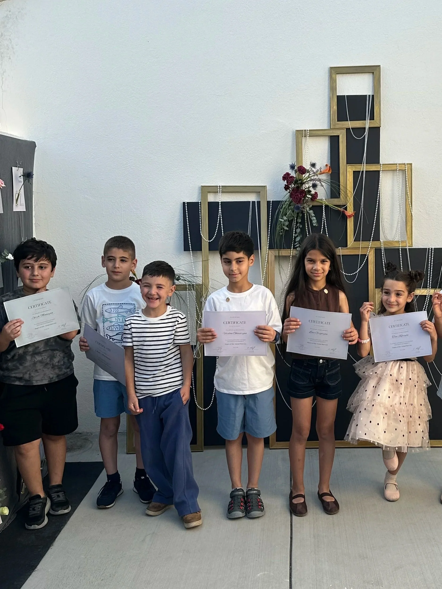 Group of six children standing on a stage, holding certificates, with a decorative display of empty picture frames, flowers, and pearl necklaces behind them.