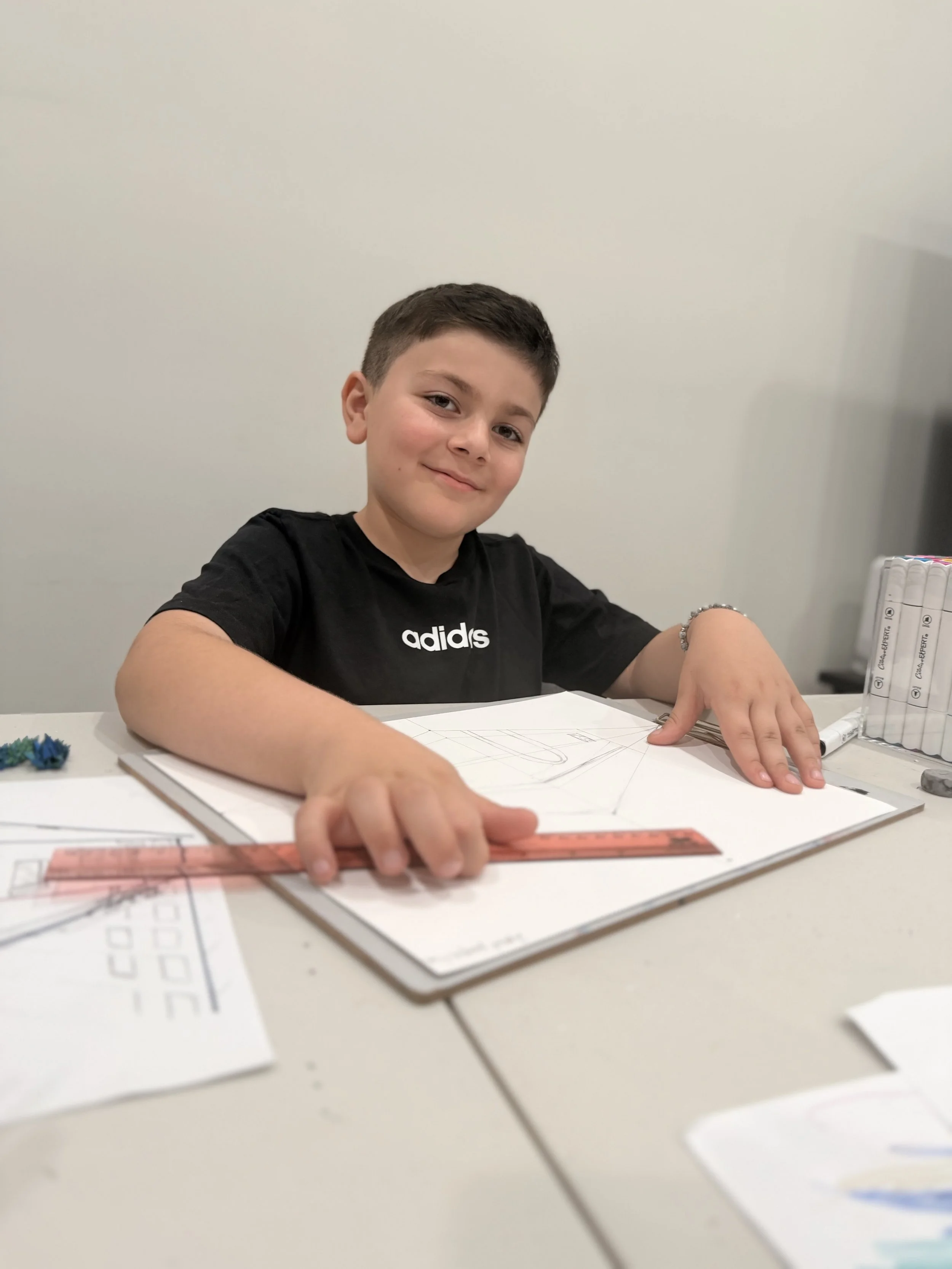 A young boy with short dark hair sitting at a table, smiling at the camera. He is wearing a black Adidas t-shirt and appears to be working on a drawing or school project with papers and a ruler in front of him.