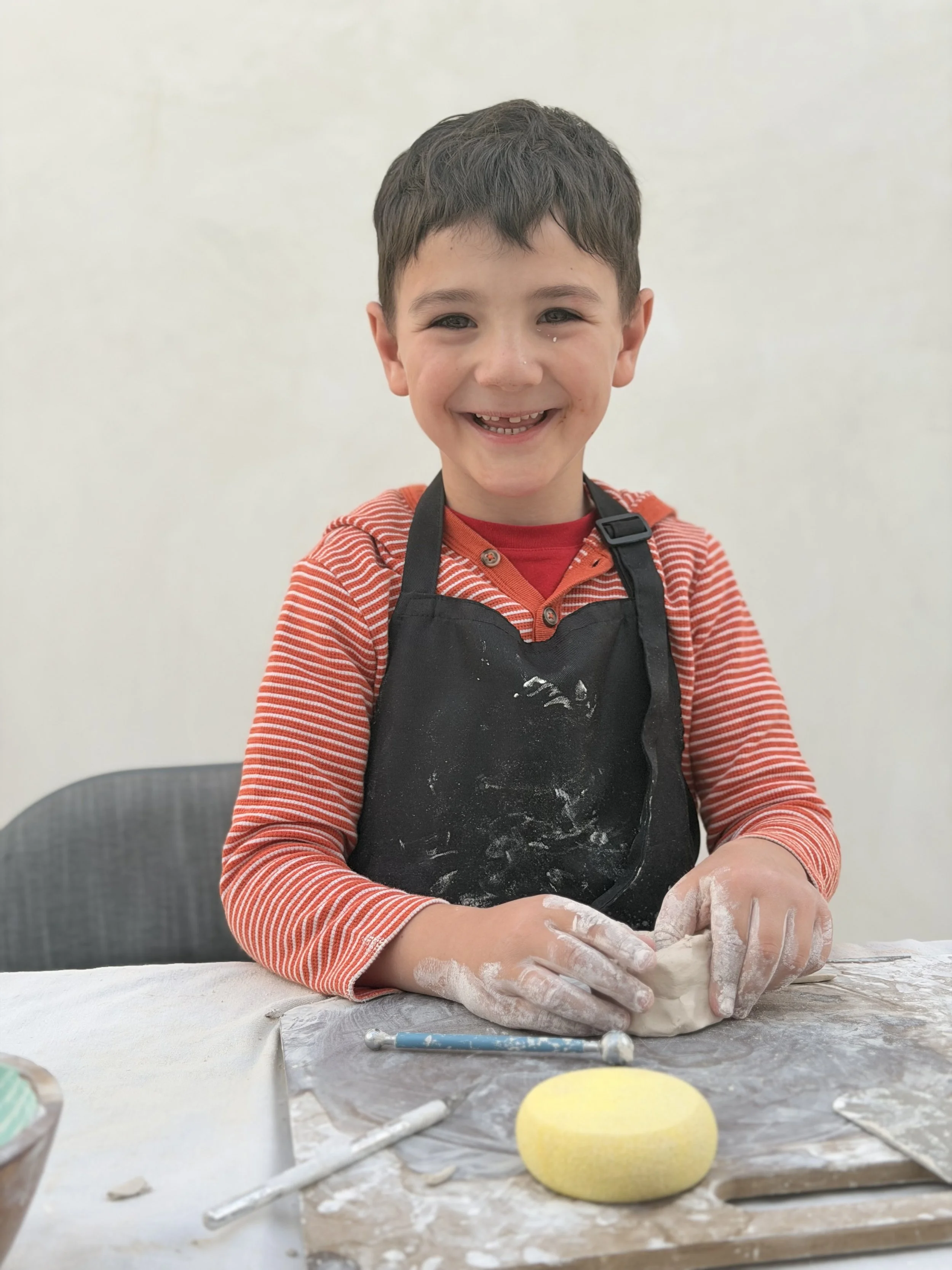 A young boy smiling while kneading dough, wearing a red and white striped shirt and a black apron, in a pottery setting.