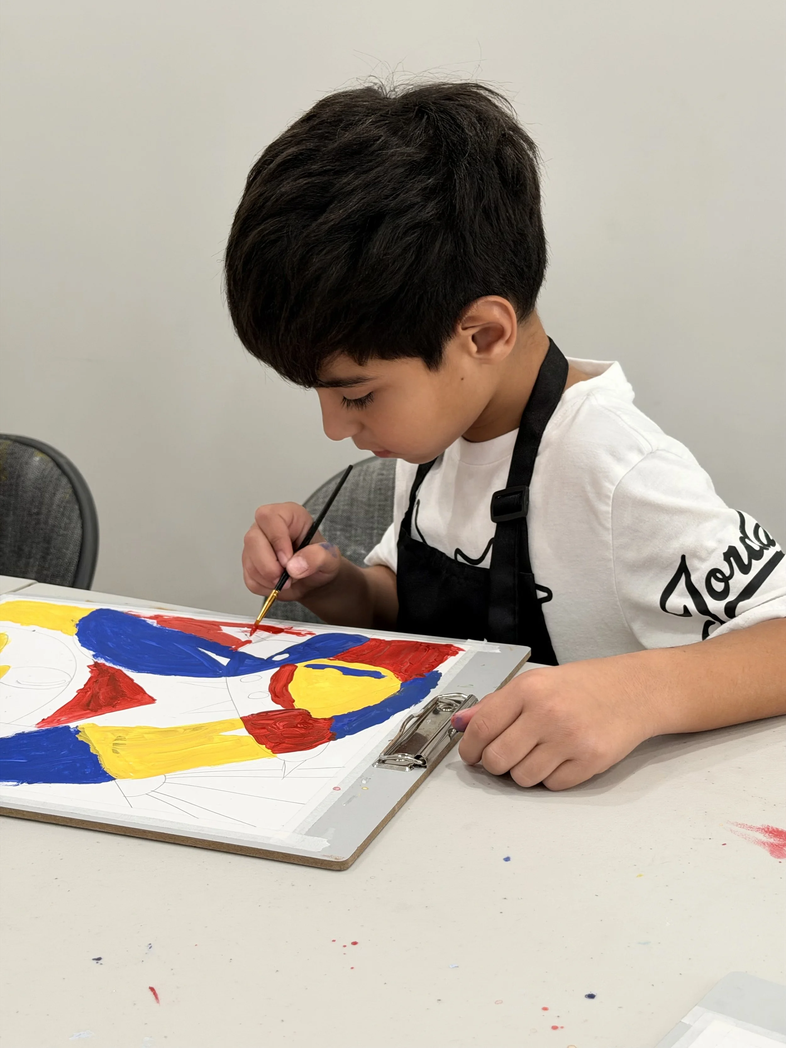 A young boy with short black hair is painting a picture of a colorful flag on a white sheet of paper. The flag has bold red, yellow, and blue sections. The boy is focused on his work, holding a small paintbrush, and is wearing a black apron over a white T-shirt.