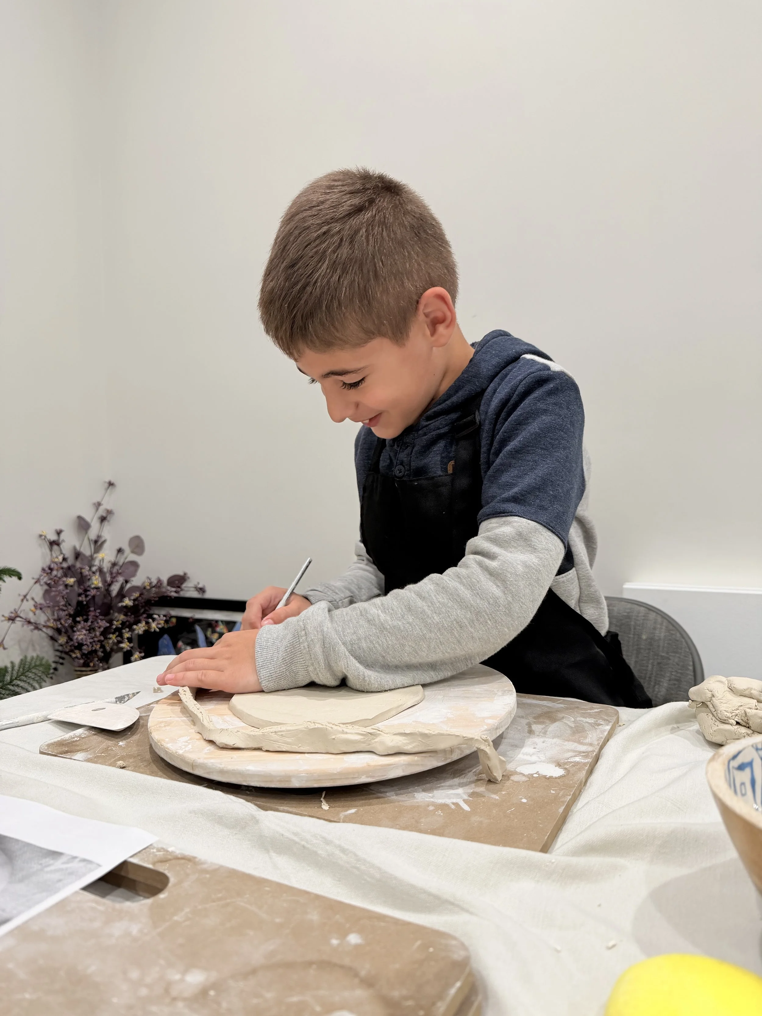A boy smiling as he works on a pottery project, sitting at a table with pottery tools and clay. There are also floral decorations next to him.