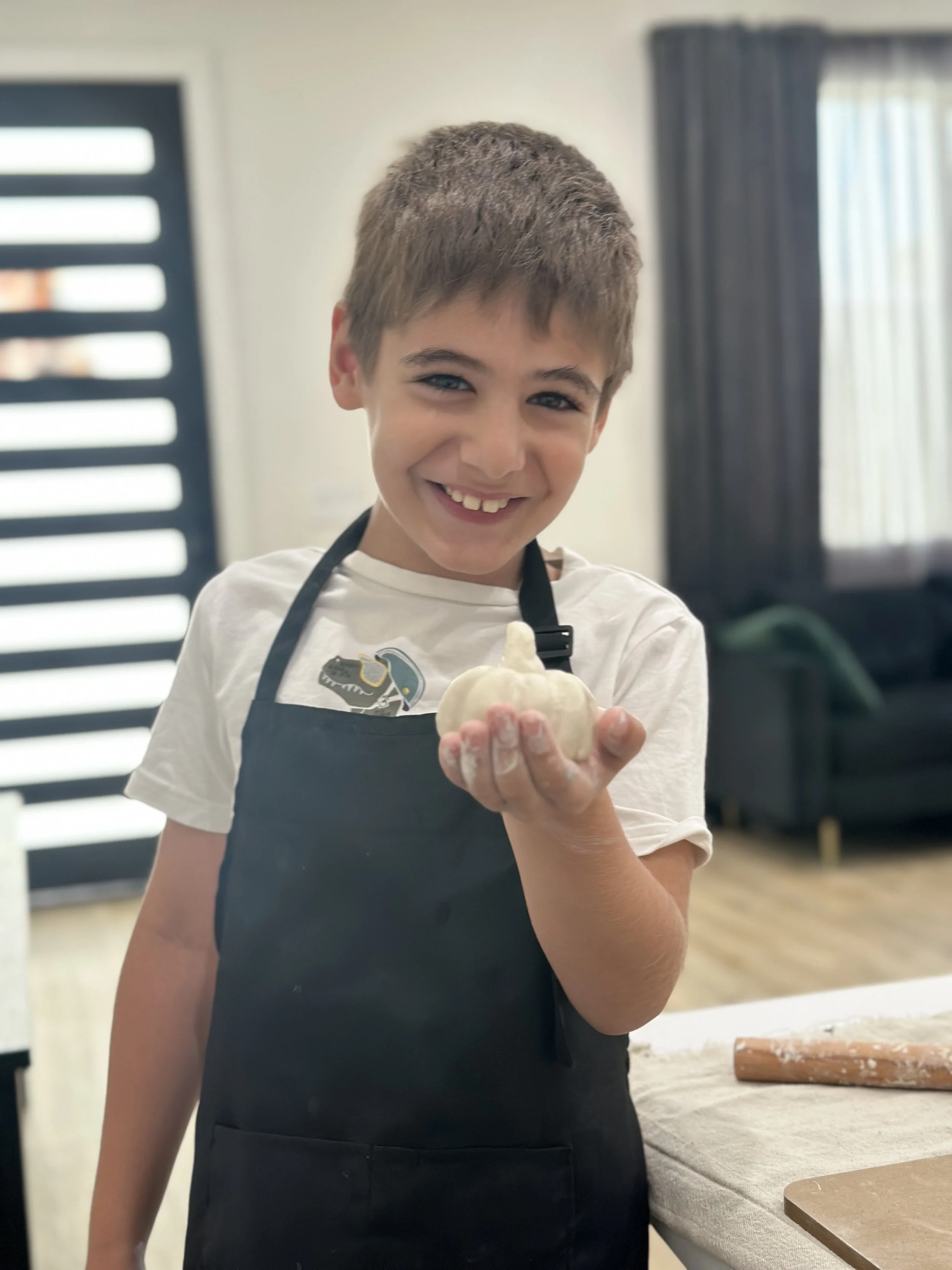 A young boy with short brown hair and blue eyes smiling while holding a piece of dough in a kitchen or baking area. He is wearing a white t-shirt and a black apron.
