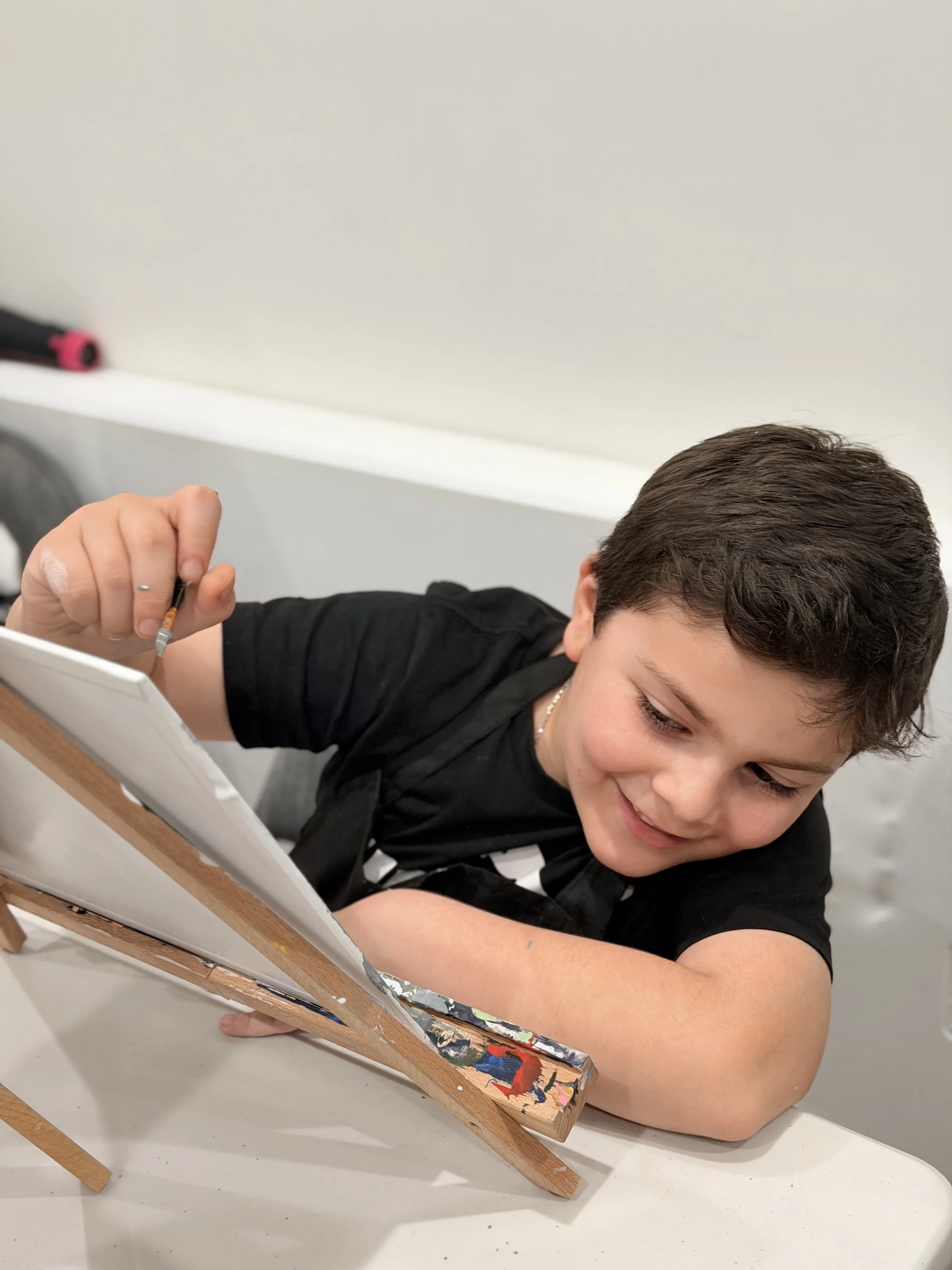 Young boy with brown hair lying on his side, smiling, leaning on his arm with an easel in front of him, painting colorful abstract art.