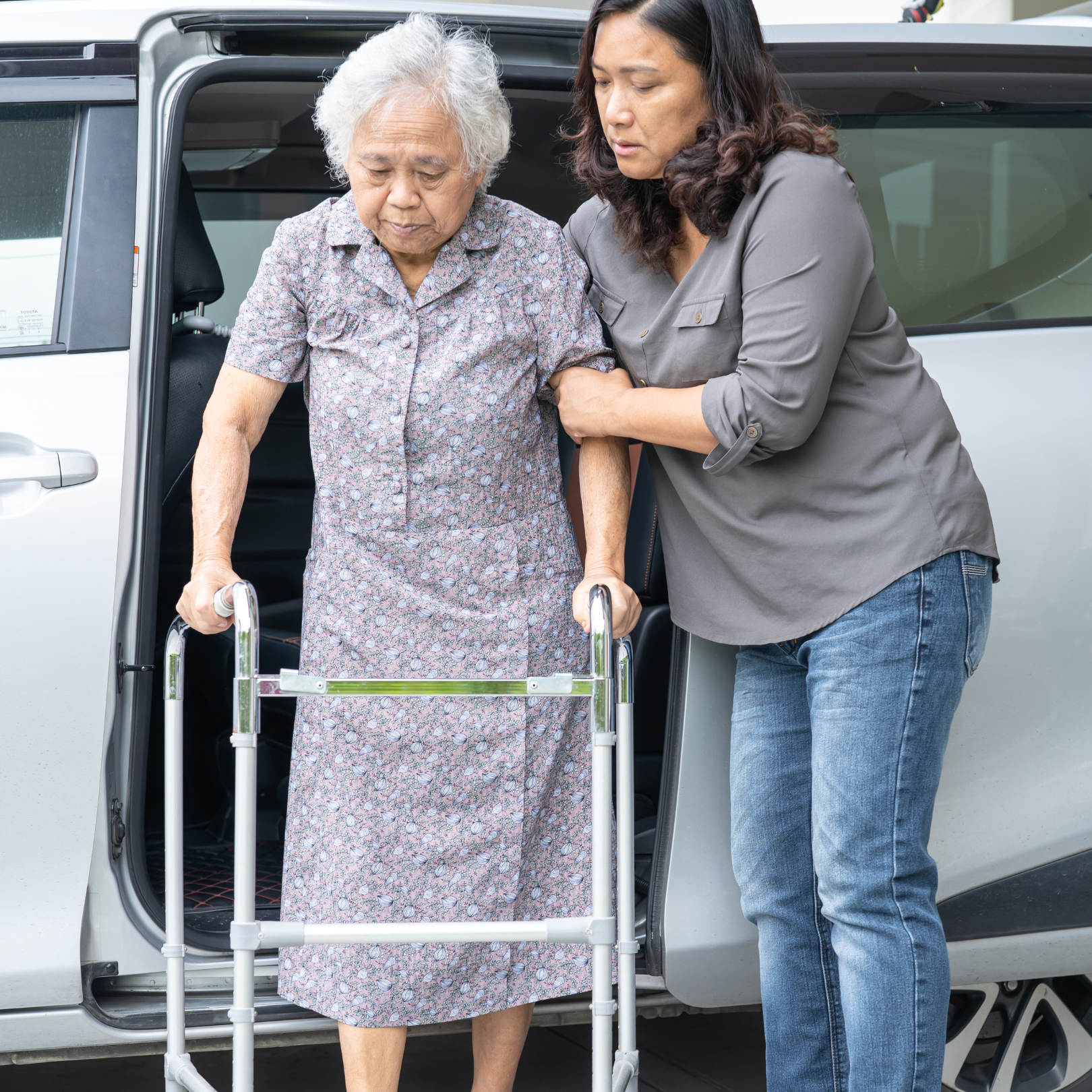 An elderly woman with gray hair using a walker being helped out of a car by a woman in a gray shirt, holding her arm to steady her.