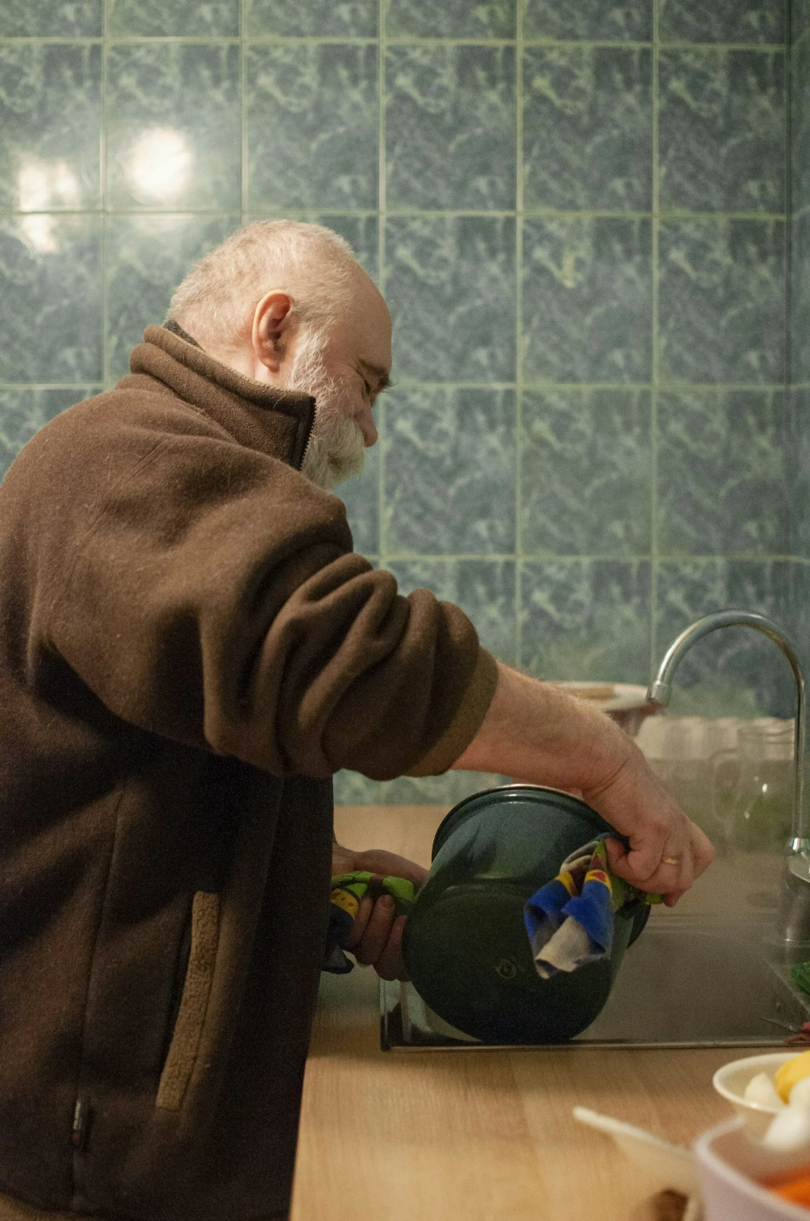 An elderly man with a gray beard and balding head washing dishes in a kitchen with green tiled walls.