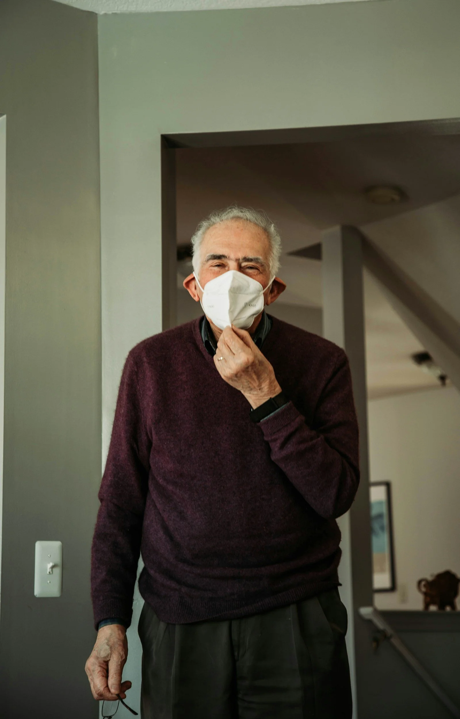 An elderly man wearing a white face mask and a maroon sweater standing indoors.