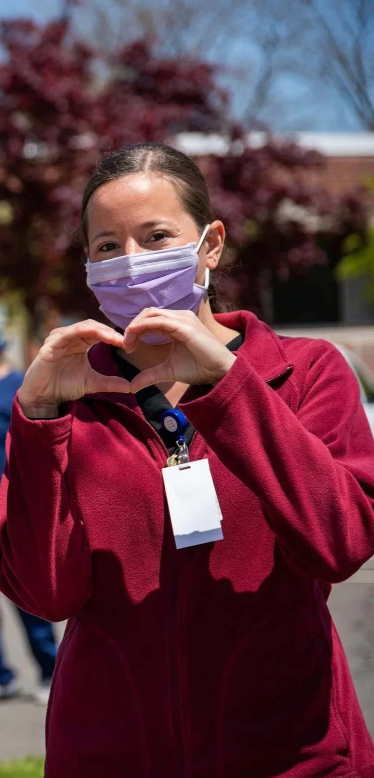 A woman wearing a purple face mask and a red jacket making a heart gesture with her hands outdoors on a sunny day.